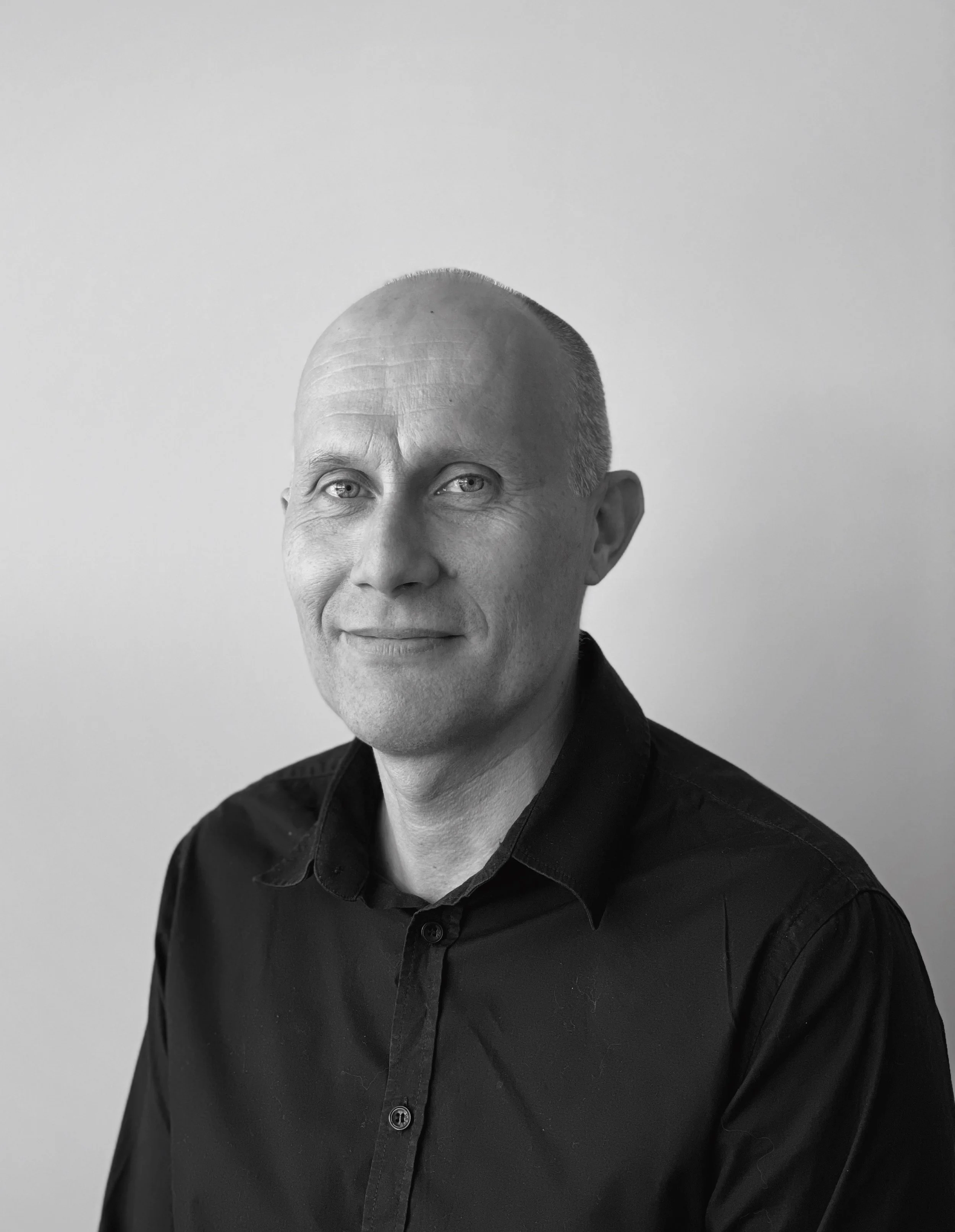 Et svart-hvitt portrett av en mann med kort hår, iført en svart skjorte, som smiler lett.