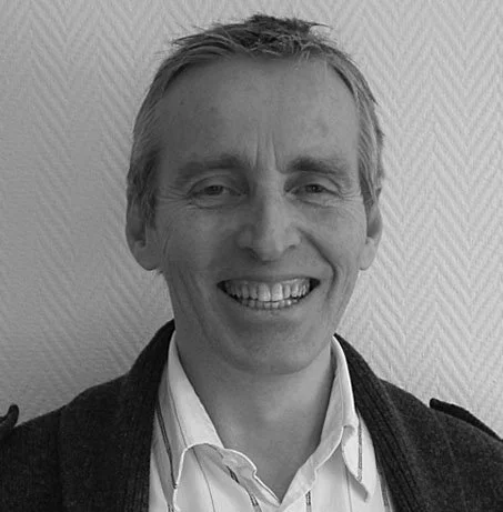 Portrett av en mann med kort, lyst hår som smiler, iført en hvit skjorte og svart jakke, mot en teksturell vegg.