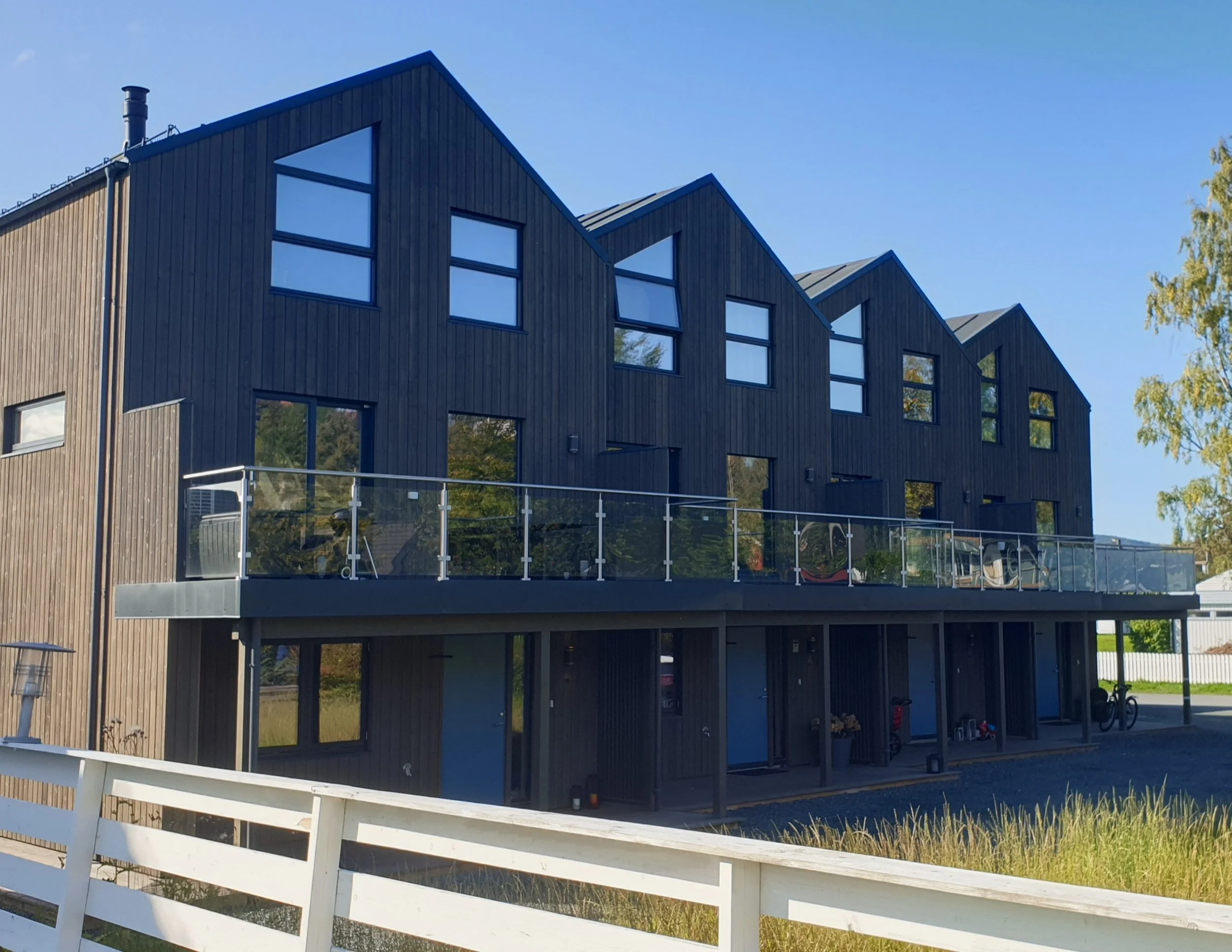 Fleretasjet fleretasjes moderne boligblokk med mørk trekledning og store vinduer, balkong med glassrekkverk, parkering under byggingen, og en hvit trehytte i forgrunnen. Solrikt og blå himmel.