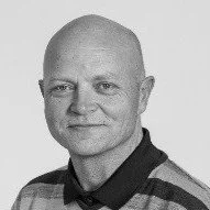 Et svart-hvitt portrett av en mann med skjegg, smilende, iført en stripete skjorte.