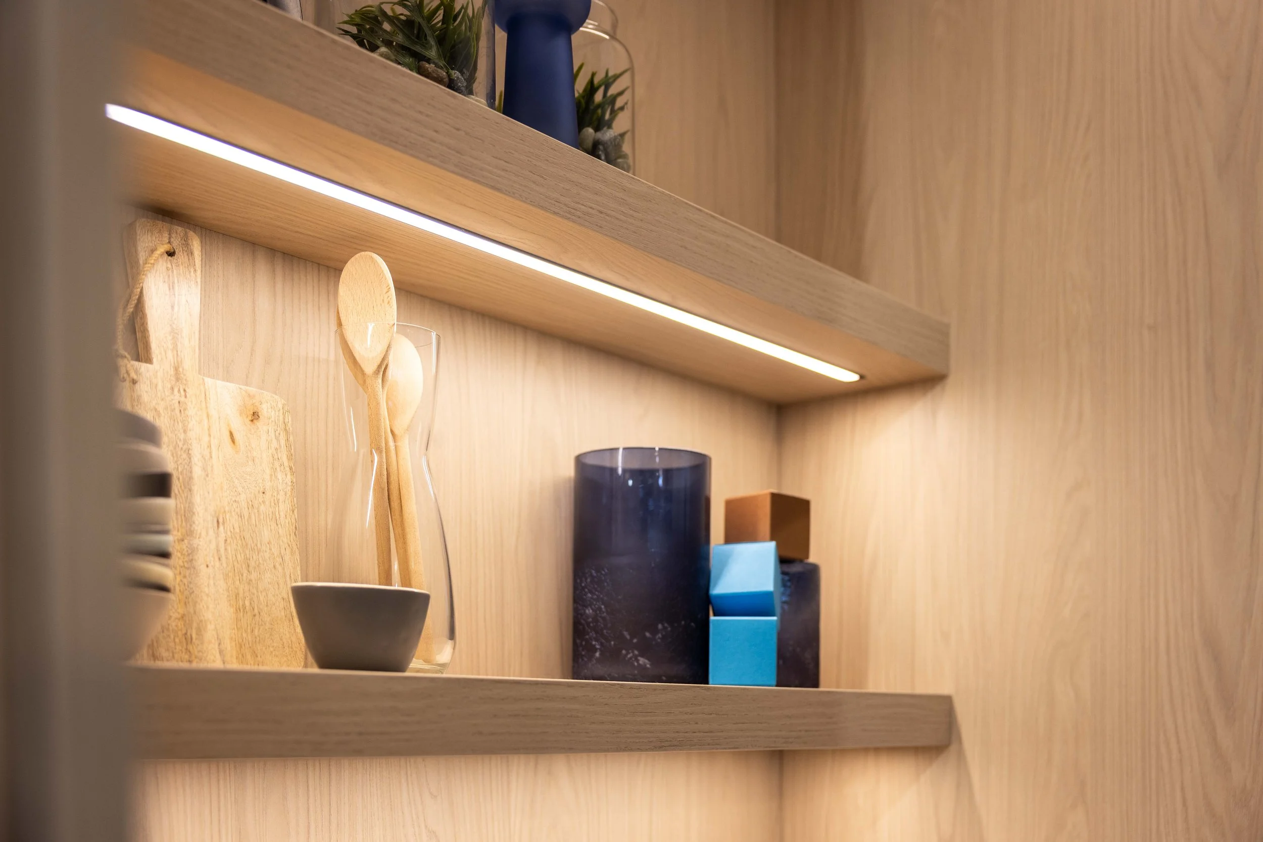 A wooden shelf with various decorative items, including a small potted plant, colorful glasses, and a blue block, with a warm wooden wall background.