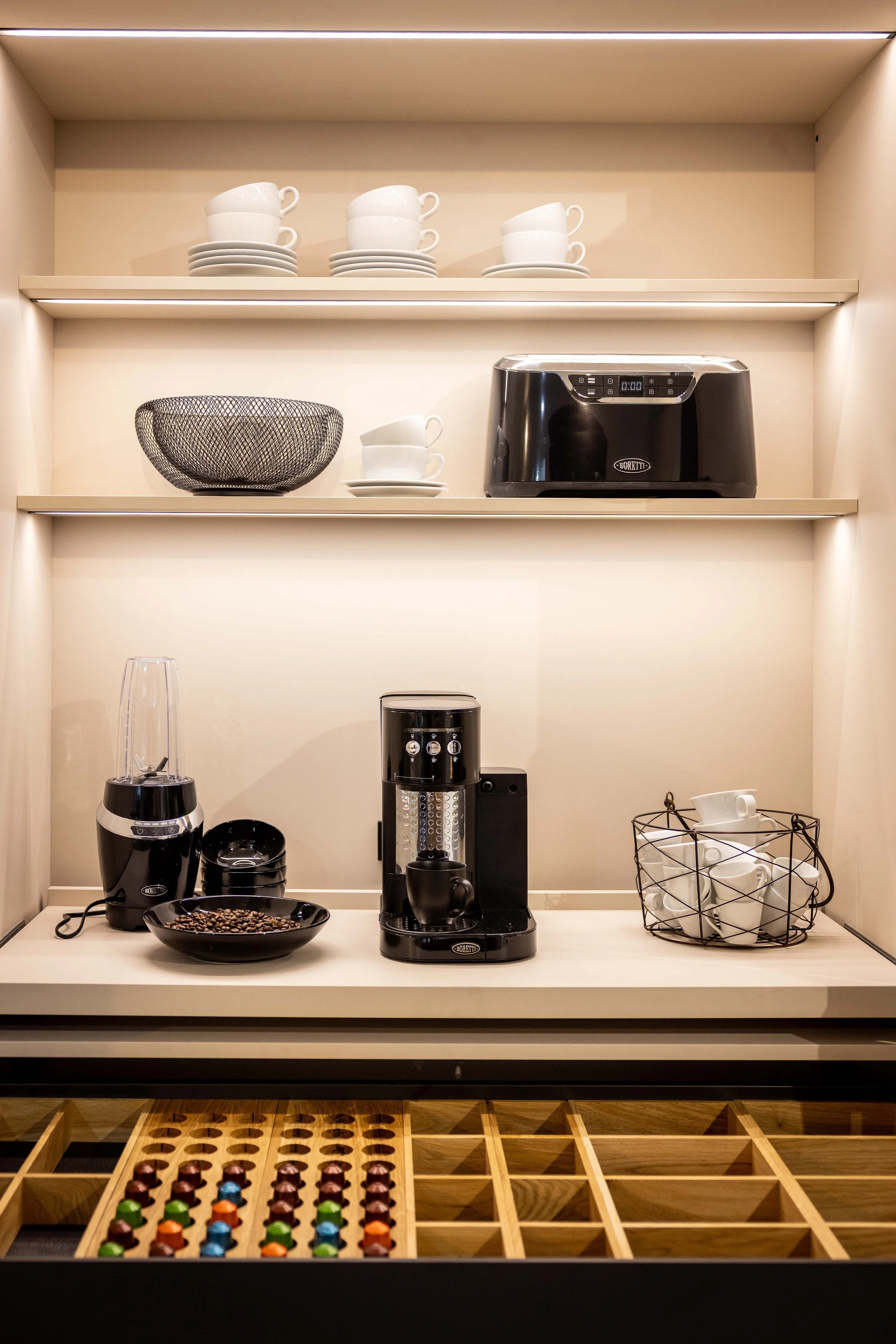 Kitchen counter with coffee grinder, coffee beans, and espresso capsules; shelves above with cups, plates, and a toaster; wire basket with white cups.