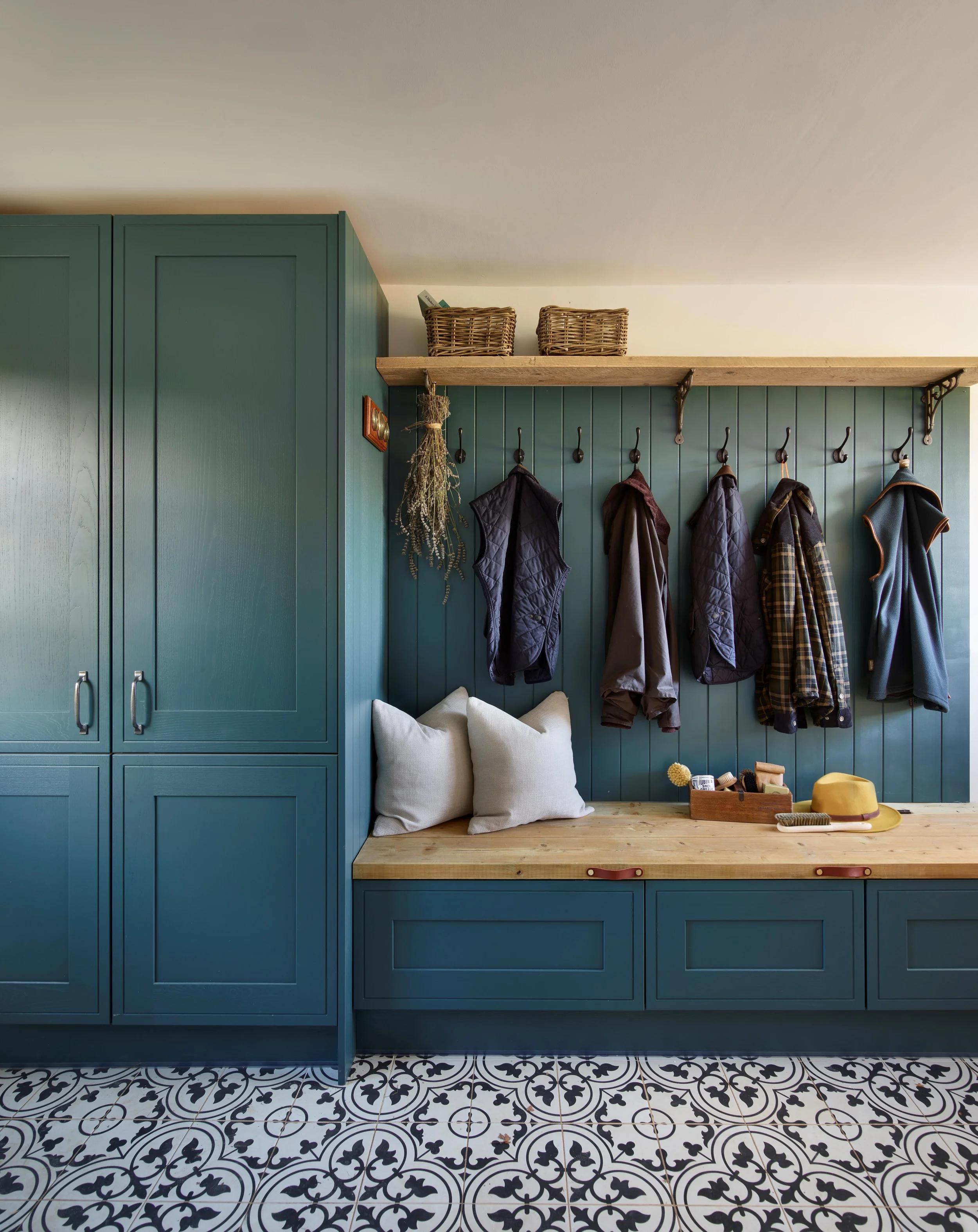 A home entryway with teal built-in cabinets, wooden bench, coat hooks holding jackets, and a patterned tile floor.