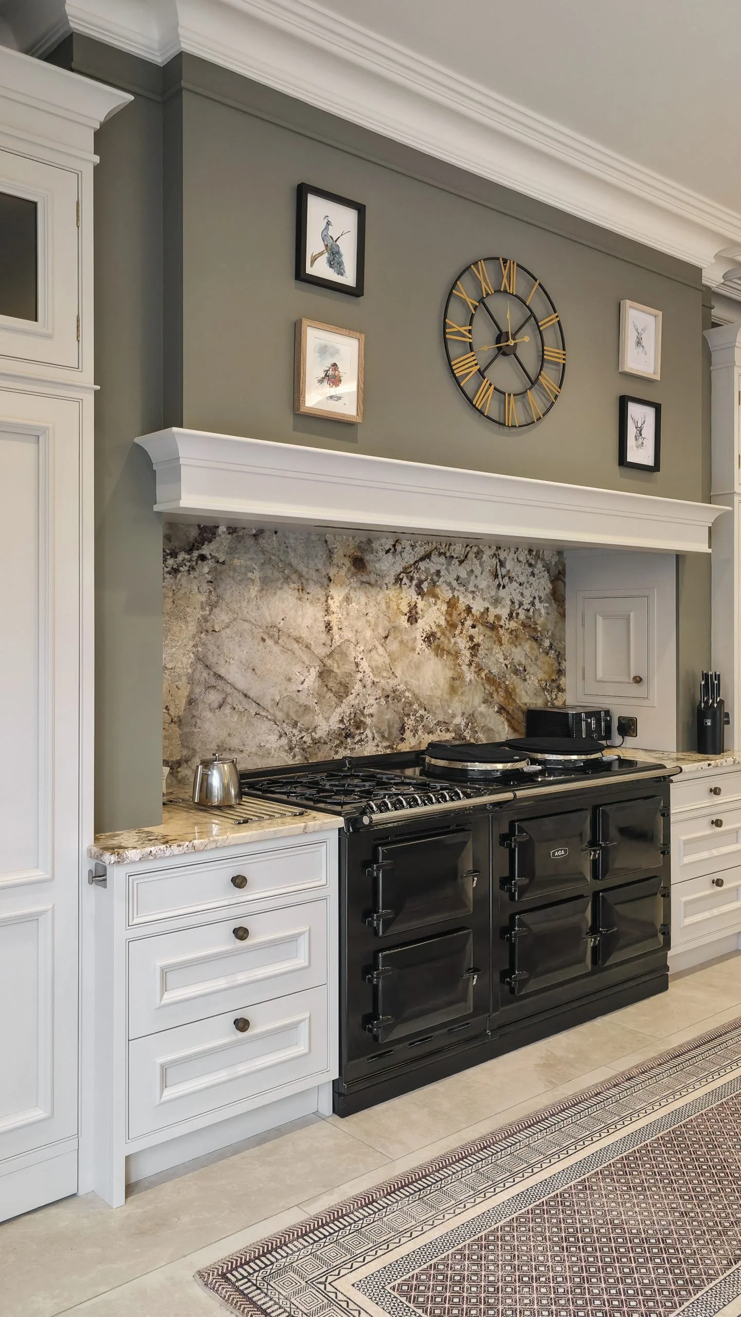Modern kitchen with white cabinetry, a black stove, a marble countertop, a stone backsplash, a clock, and framed artwork on gray walls.
