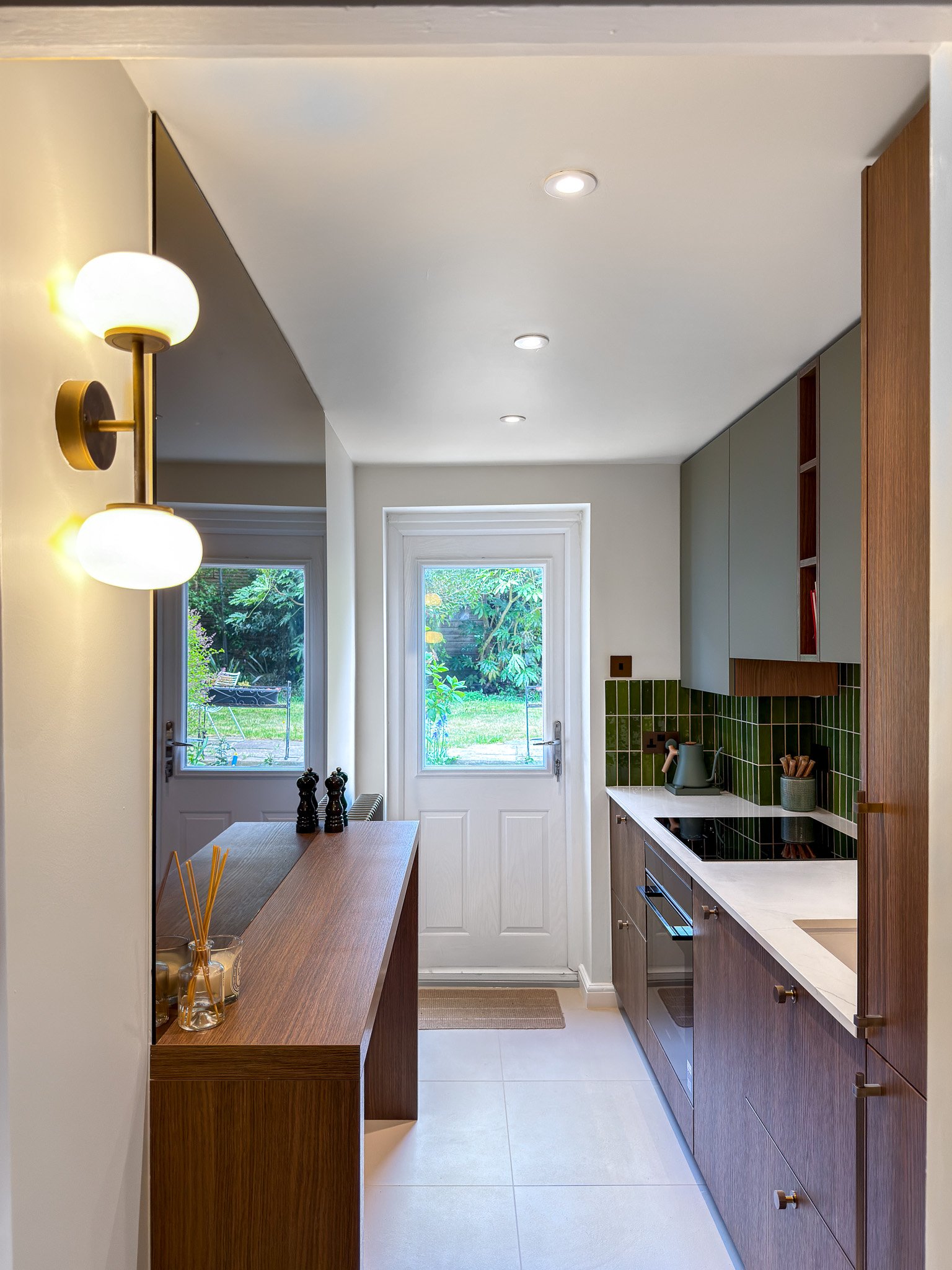 A compact kitchen with white walls, a door leading outside to a garden, and a window showing greenery. The kitchen features wooden cabinets with a white countertop, green tiled backsplash, and modern appliances. There is a wooden breakfast bar with items on it, and the room is illuminated by ceiling lights and a wall-mounted light fixture.