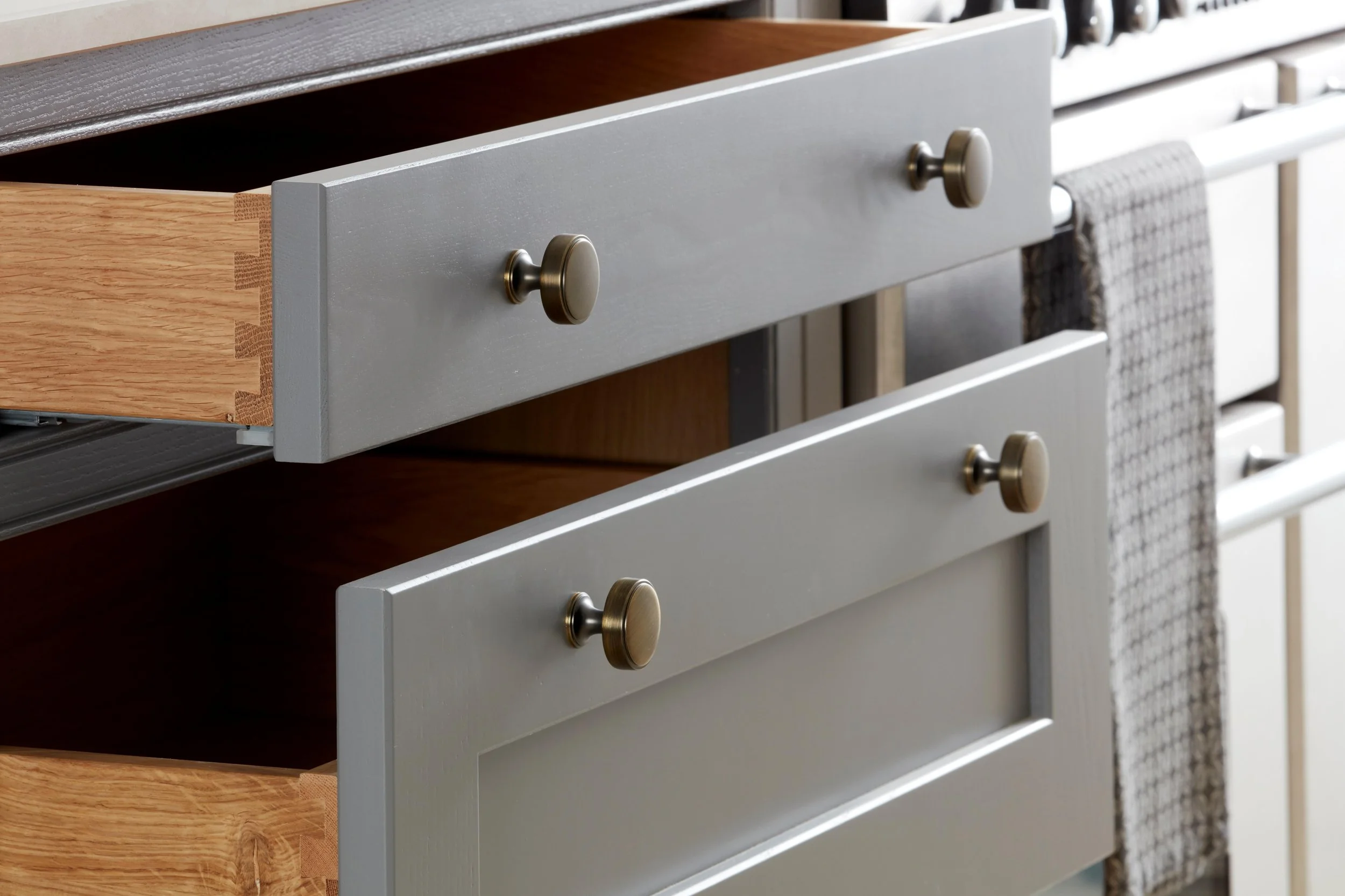 Close-up of a light gray wooden kitchen drawer with round brass handles, partially open, revealing empty interior and neighboring drawers with different finishes, including wood and fabric.