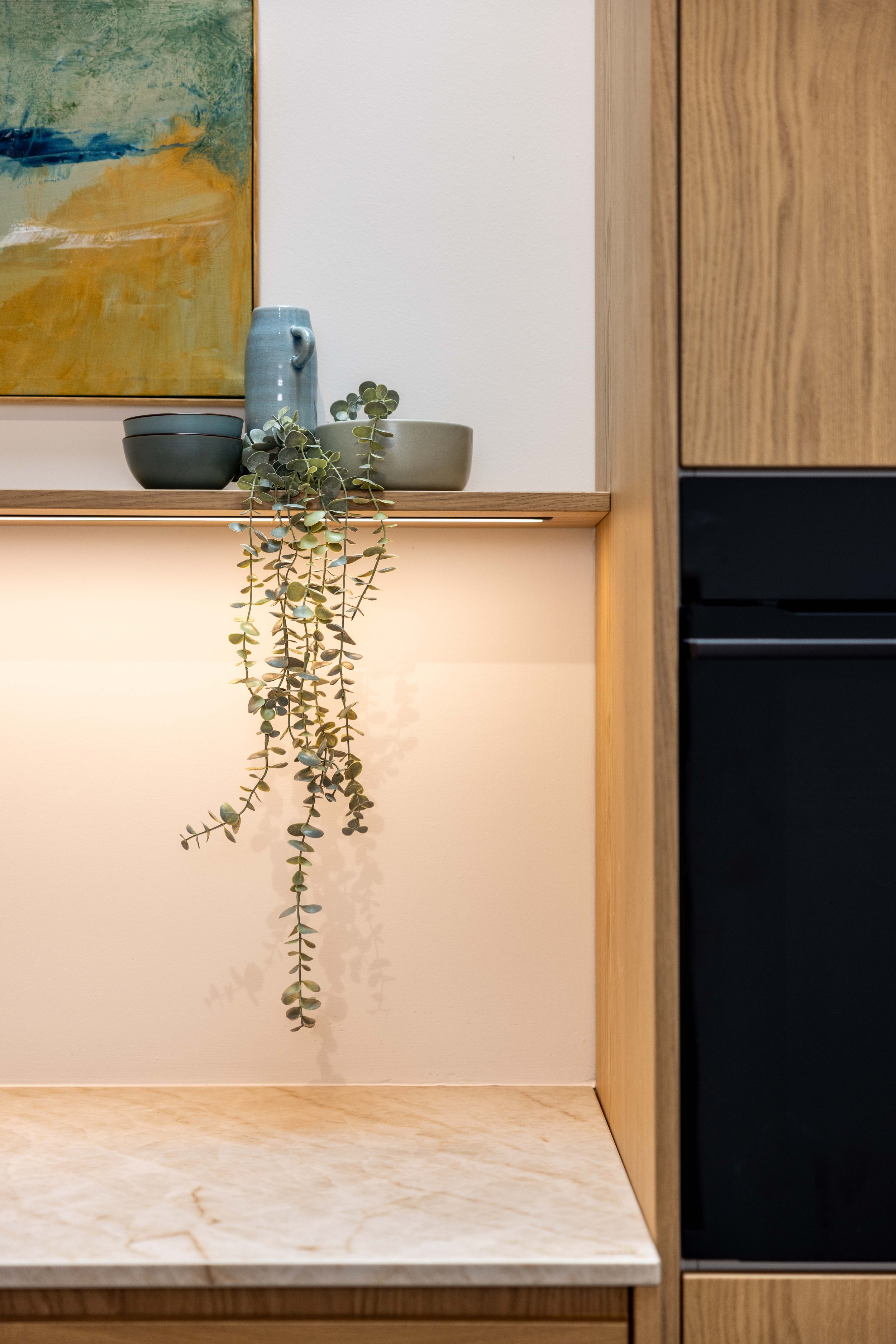 A minimalist kitchen shelf decorated with a light blue ceramic pitcher, two looping bowls, a small green eucalyptus plant, and a framed abstract painting with yellow, green, and blue tones on a white wall.