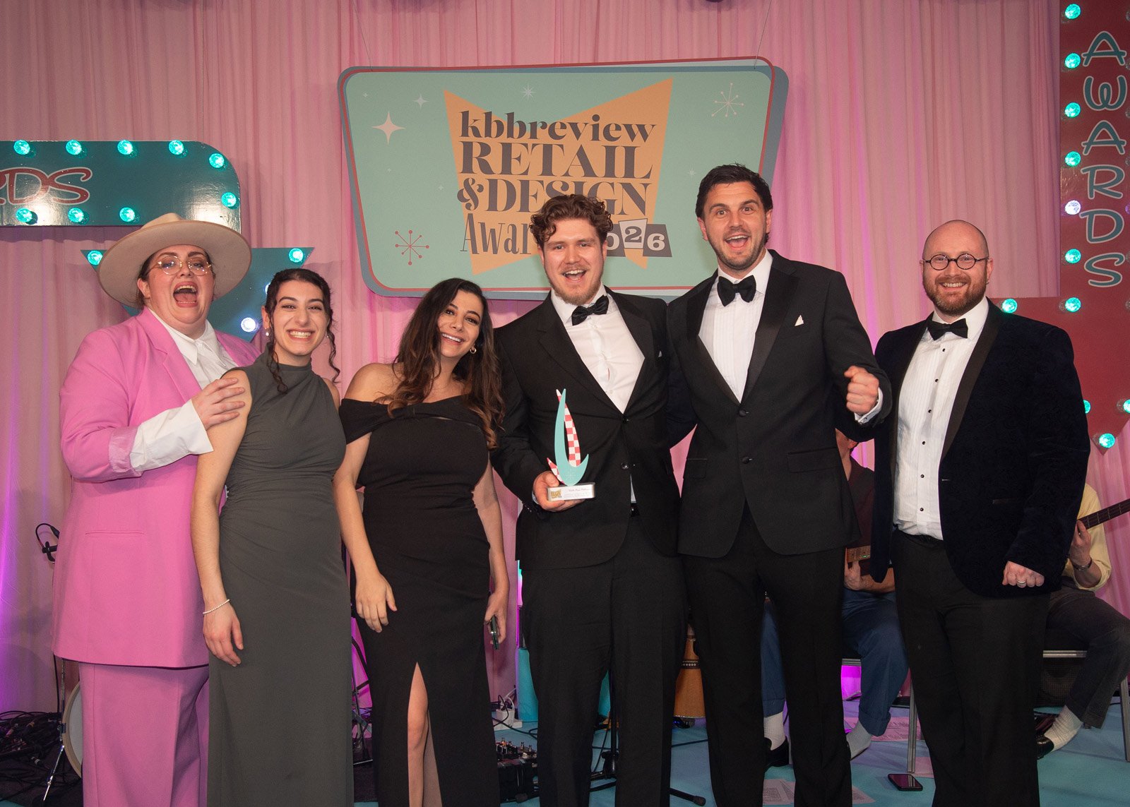 Group of six people in formal attire on stage at an awards event with a pink curtain background and a sign that reads 'Kbbrewview Retail & Design Awards 2023'.