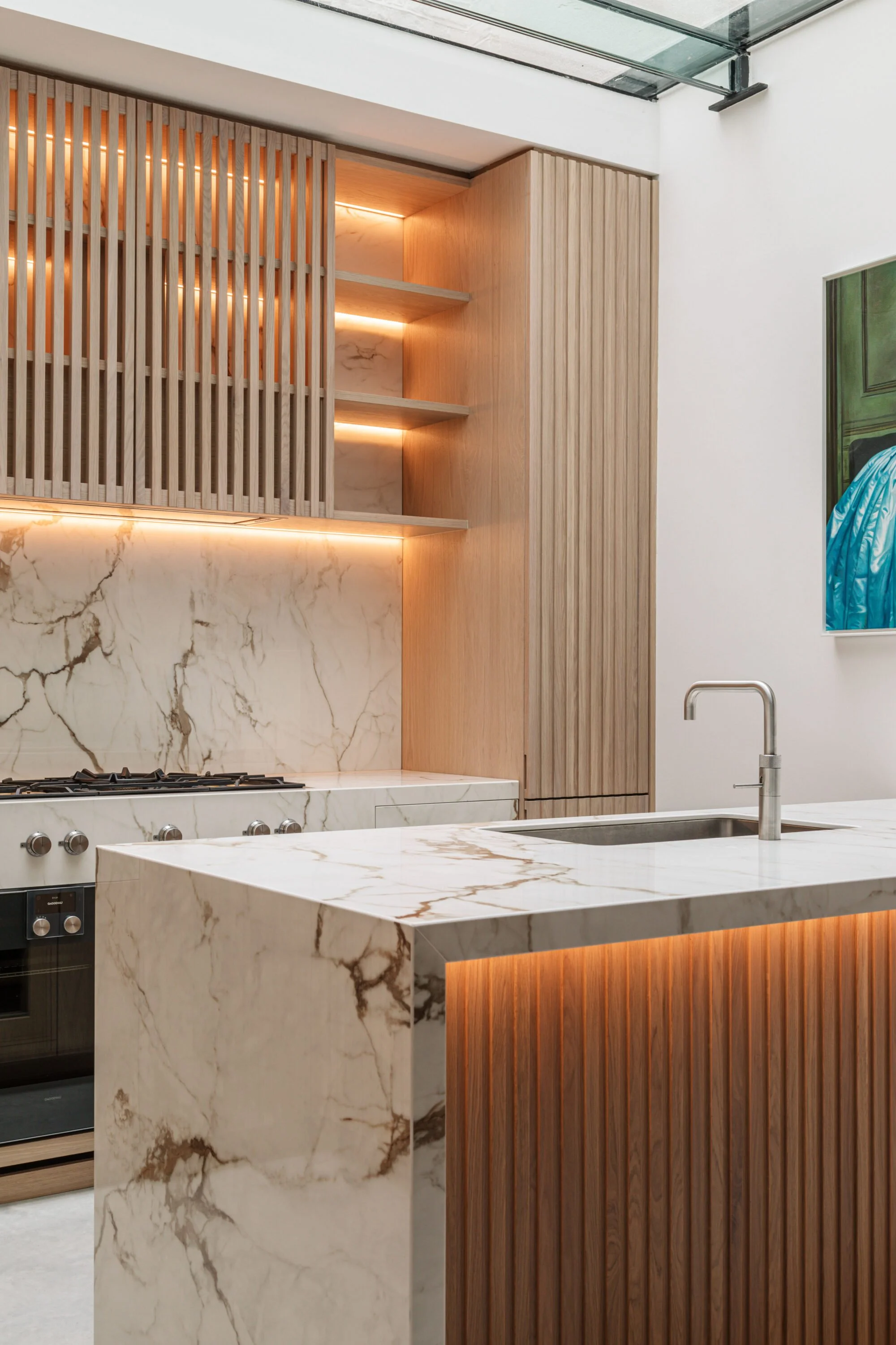 Modern kitchen with marble countertops, a sink, and wooden cabinetry with vertical slats, illuminated by soft lighting and a skylight overhead.