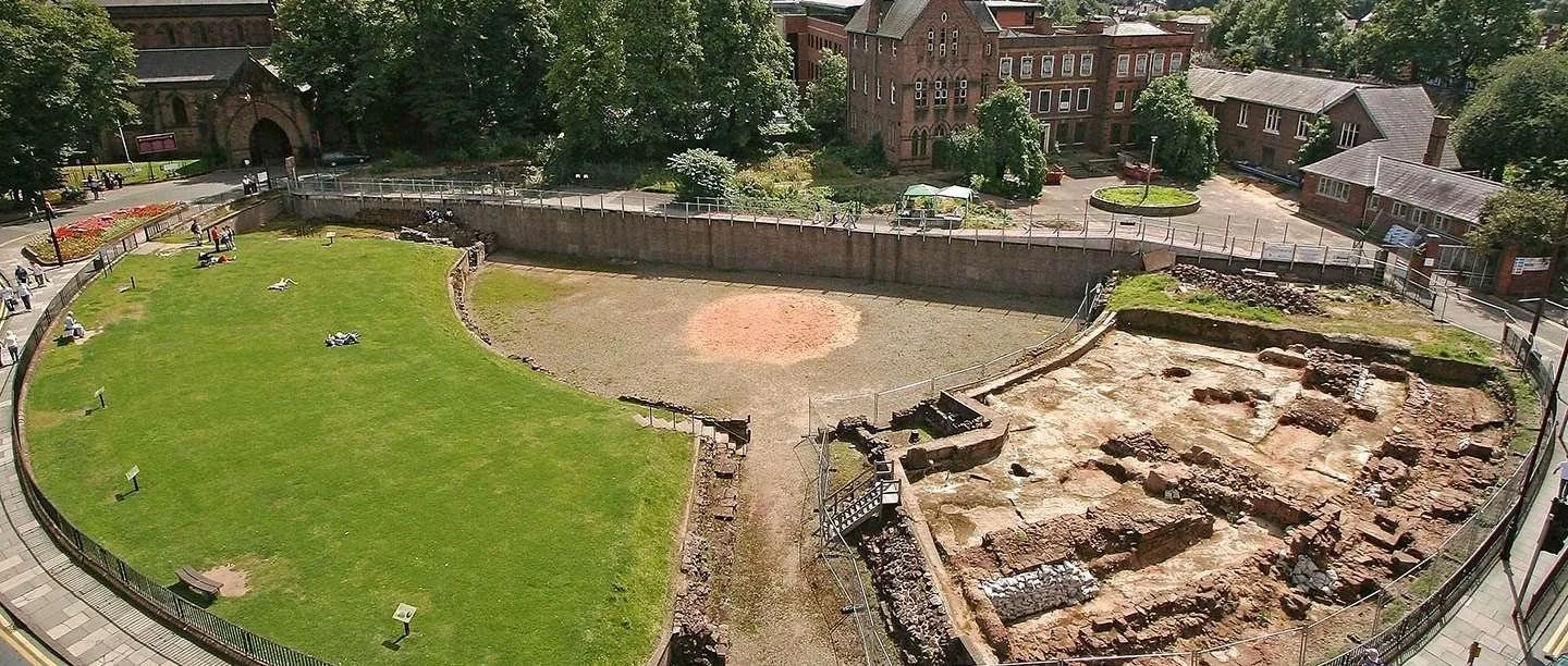 An aerial view of an archaeological site with ancient ruins, adjacent to a green lawn and surrounded by a sidewalk in a city neighborhood.