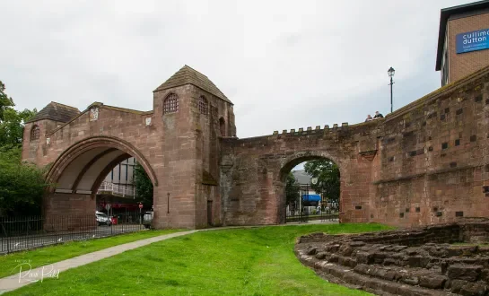 A historic stone fortress with arched entryways, surrounded by a grassy park and a paved walkway, with modern buildings and trees in the background.