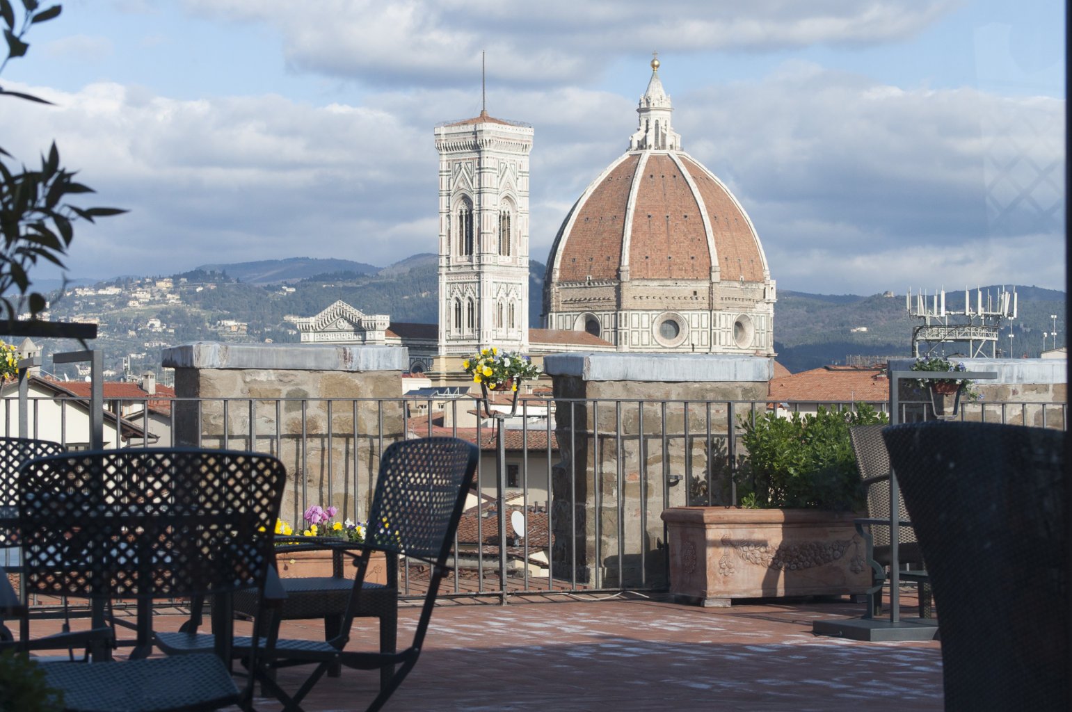 Rooftop terrace view of Florence, Italy, featuring the Cathedral of Santa Maria del Fiore with its iconic red-tiled dome and the adjacent Bell Tower, with outdoor furniture and potted plants in the foreground.