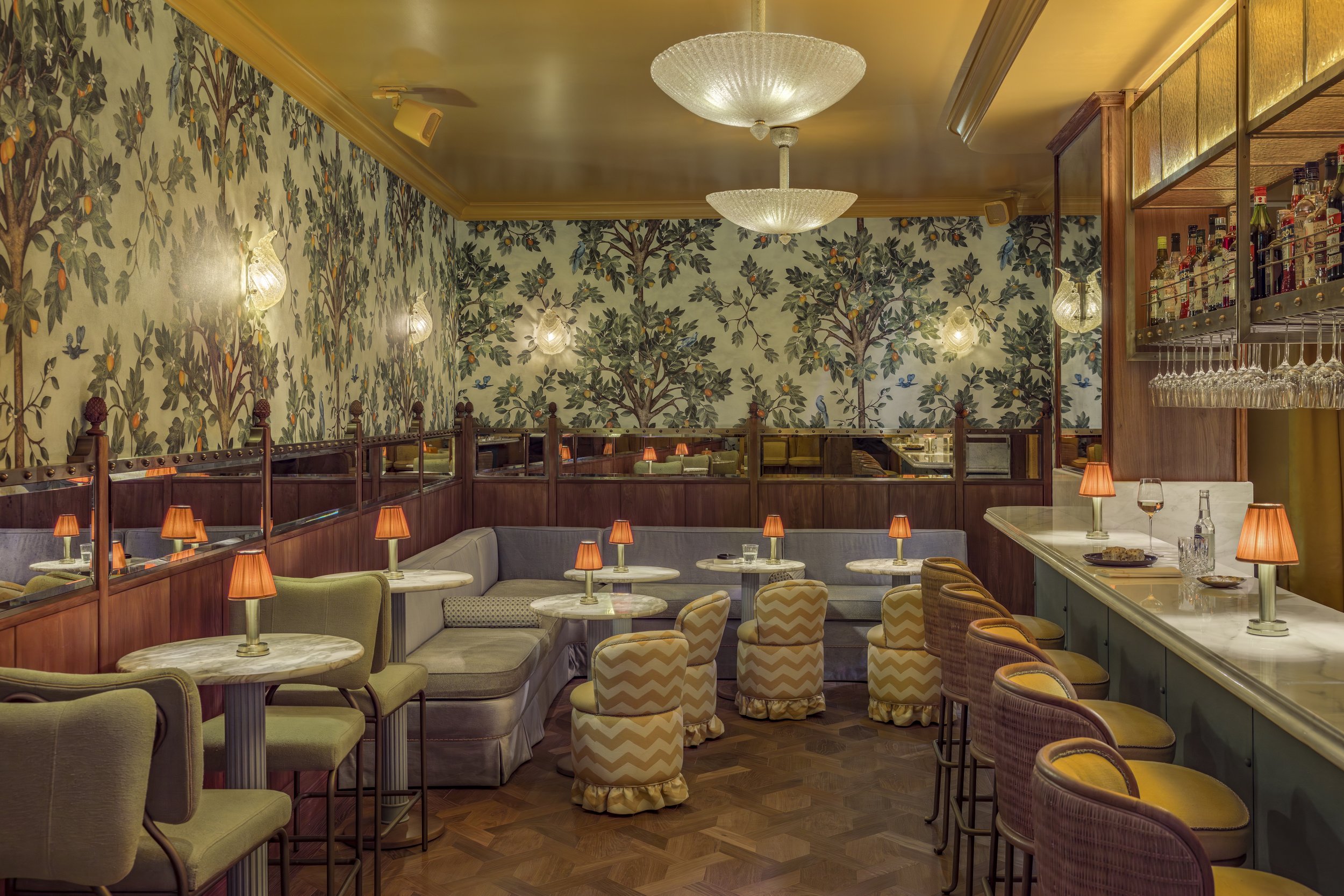 La Fantaisie Mumbai India Cafe with golden ceiling lamps, Adam Ellis tree-and-bird wallpaper, pastel green and cornflower blue velvet seating, parquet floors, and heavy mustard drapes in maximalist dining interior