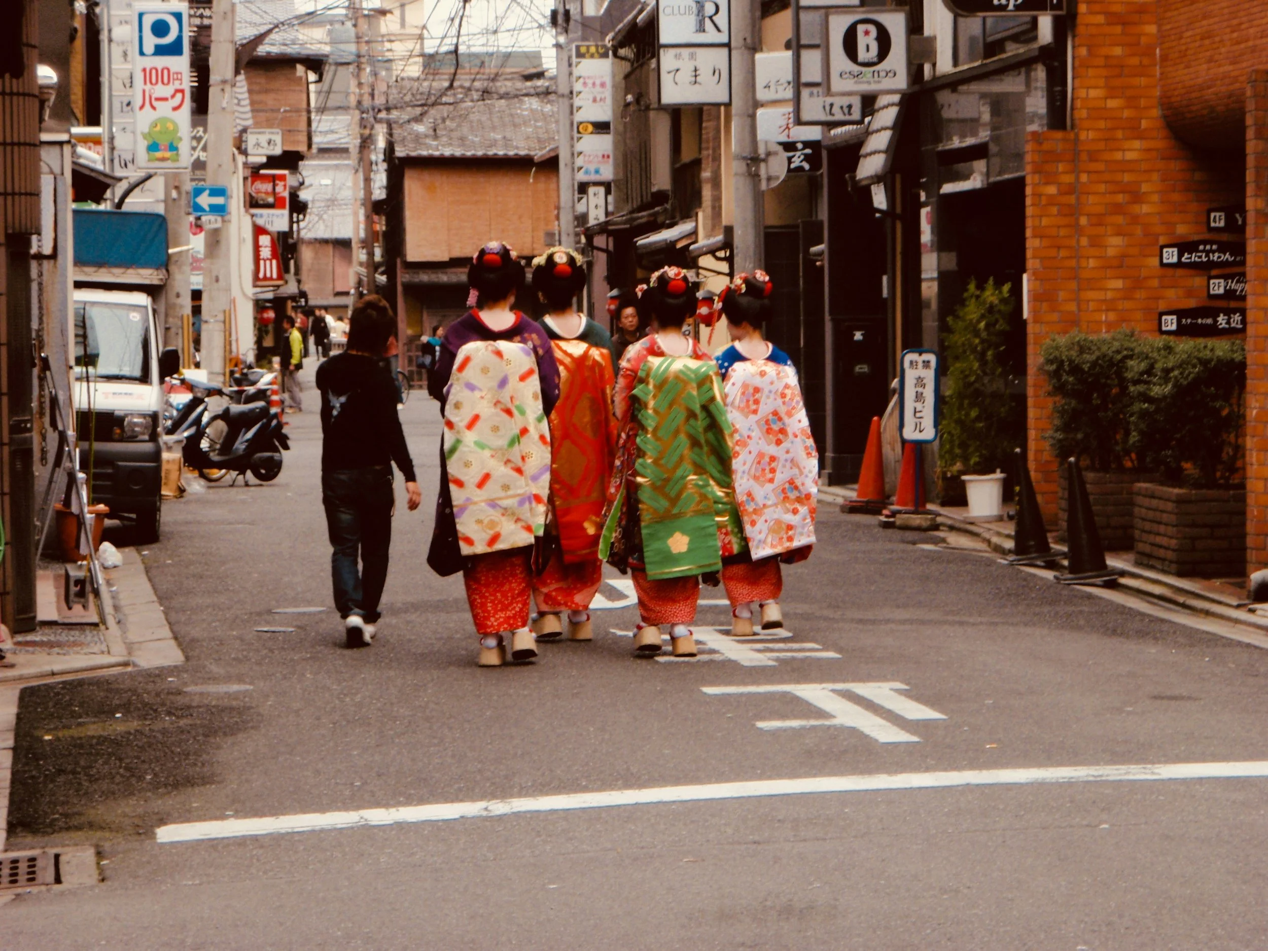 A group of four women dressed in traditional kimonos walking down a street in Japan, seen from behind.