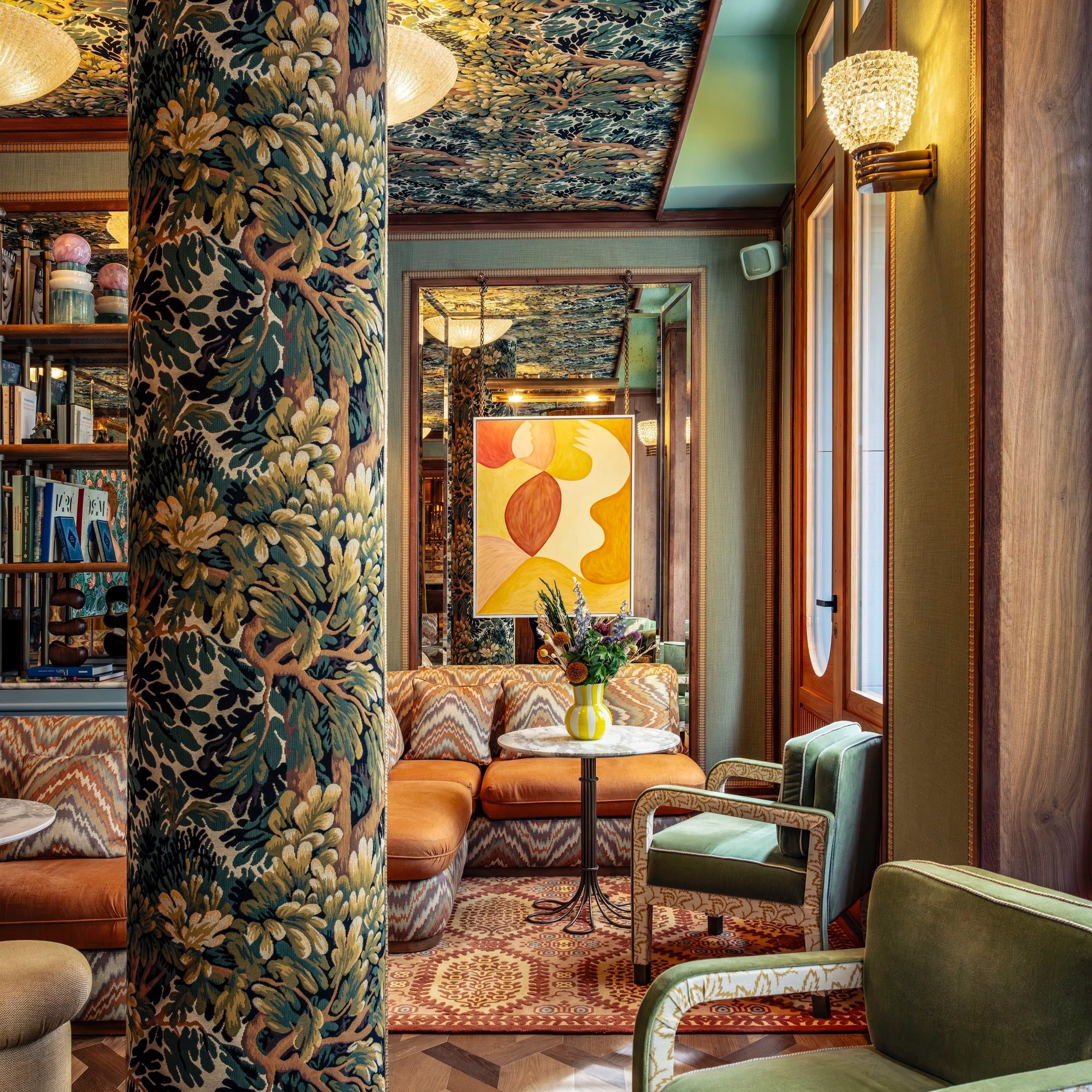 La Fantaisie Mumbai India lobby with maximalist interior design, foliage-printed central column, mirrored walls, burnt-orange chevron banquettes, green armchairs, and dense ceiling of leaves and branches in theatrical, indulgent decor