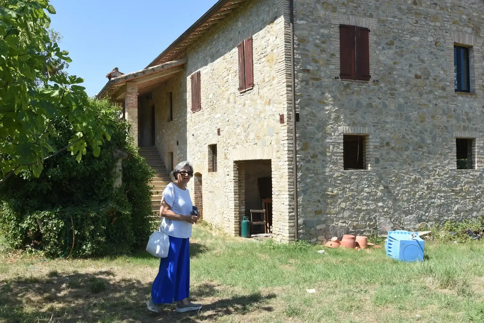 Elvira walking on green lawn before a rustic Umbrian house with stairs.
