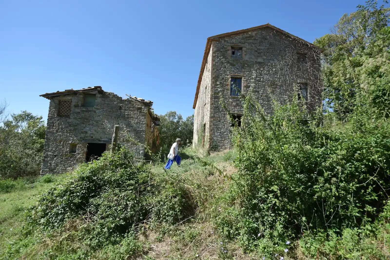 Elvira walking between two rural Umbrian houses under a clear blue sky.