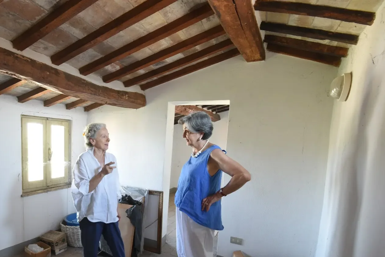Two women talking inside a rustic room with wooden ceiling and natural light.
