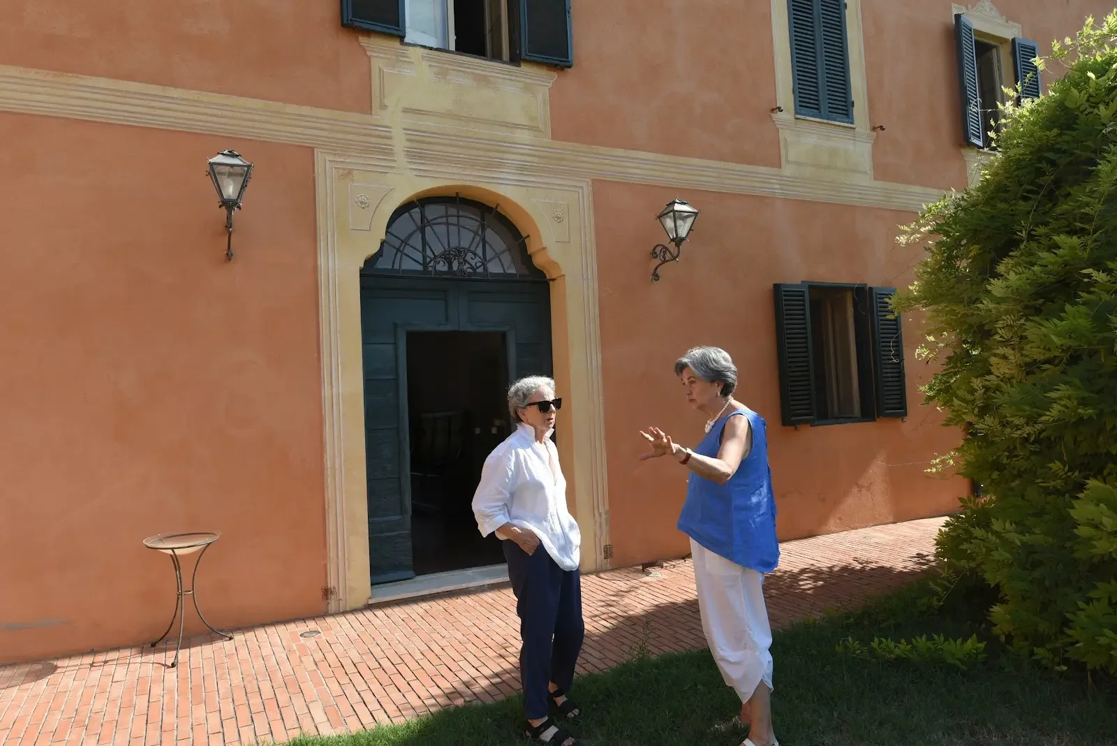 Elvira chatting with a woman outside a rustic orange house with shutters in a sunny Umbrian day.