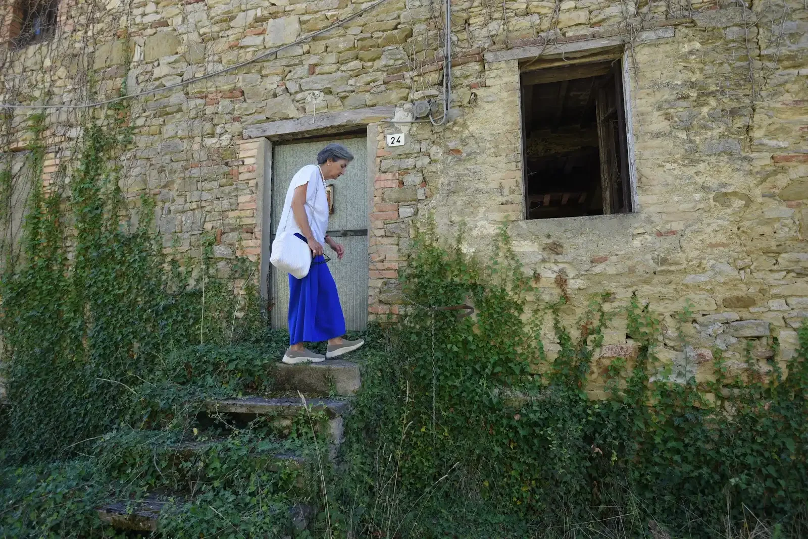 Elvira standing on stone steps before a traditional Umbrian home with climbing plants.
