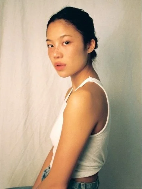 Portret van een jonge vrouw met een lichte huid en donker haar, zittend tegen een neutrale achtergrond, gekleed in een witte top.