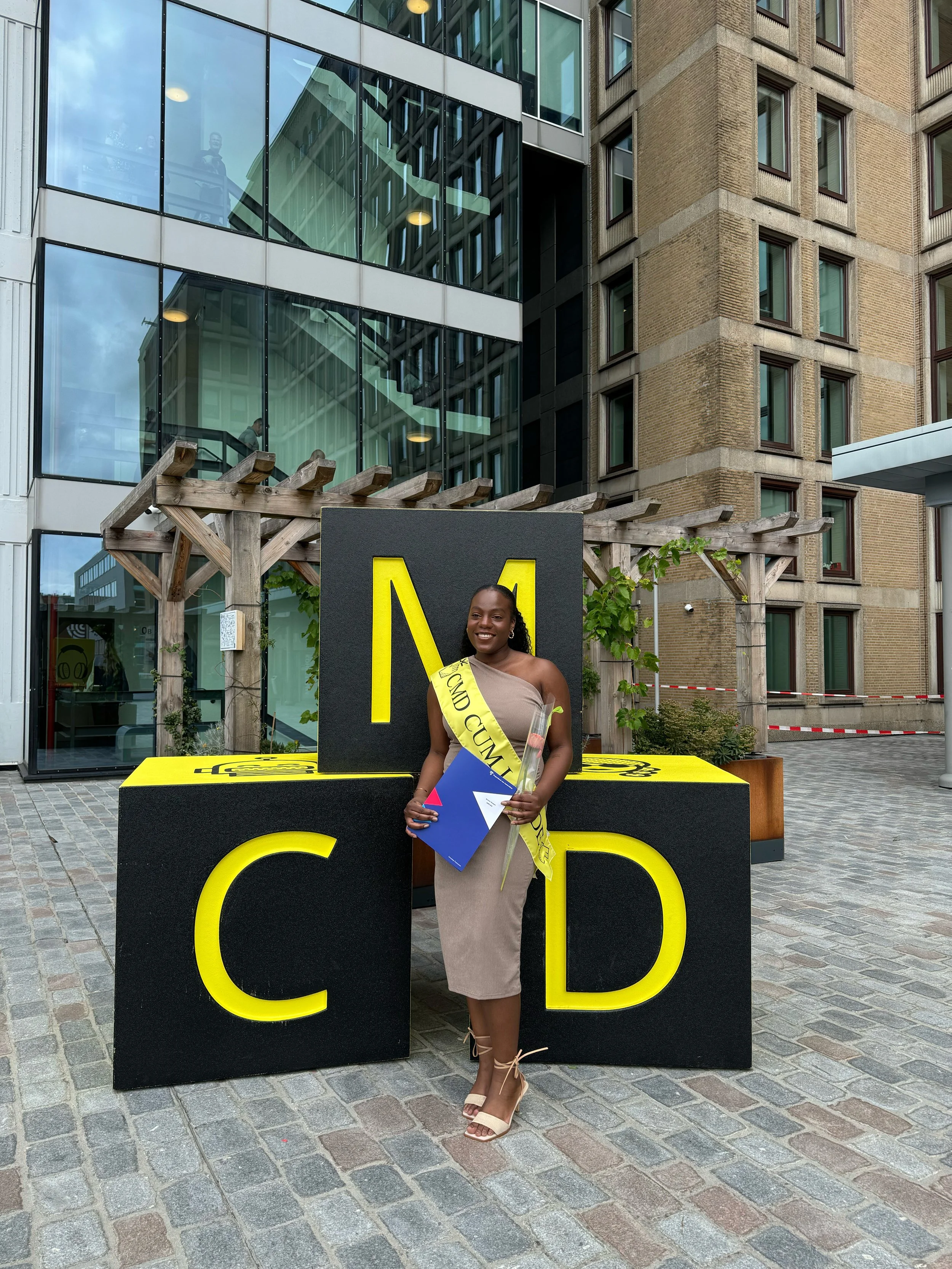 Vrouw in beige jurk met sjerp die 'BRAVO CUM LAUDE' draagt, standing voor grote letterblok 'CMD', holding bloemen en diploma in een stedelijke omgeving.
