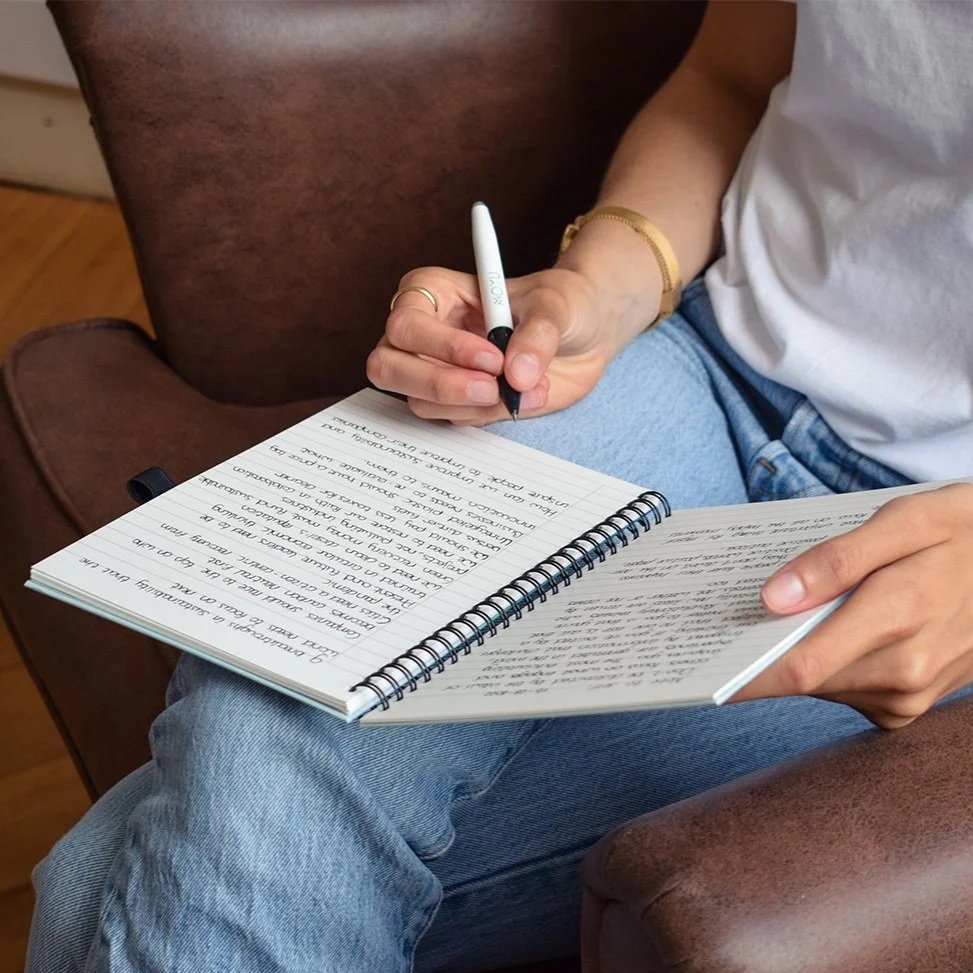 Persoon schrijft in een open ringband notitieboekje terwijl ze op een leren stoel zit.