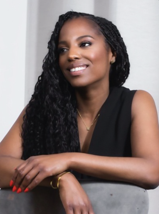 Een vrouw met lang, krullend zwart haar die glimlacht en een zwarte top draagt, zittend met haar armen op een tafel.