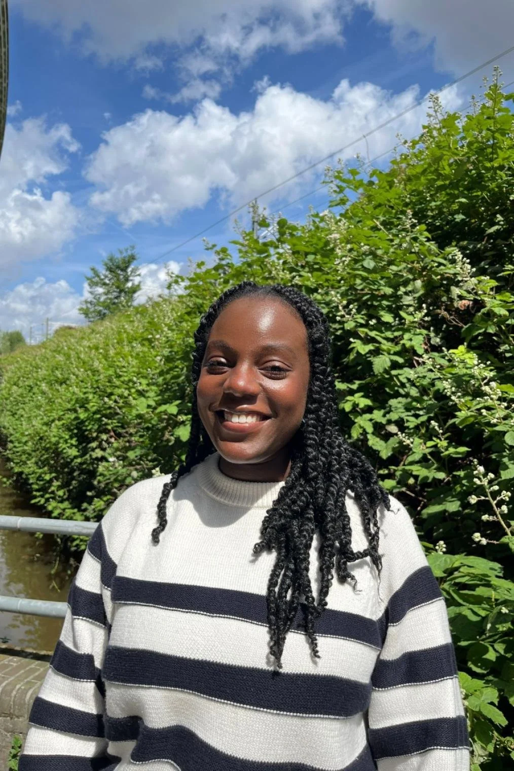 Een lachende vrouw met zwart haar in een zwart-wit gestreepte sweater, Staande voor een groene heg onder een blauwe wolkenlucht.
