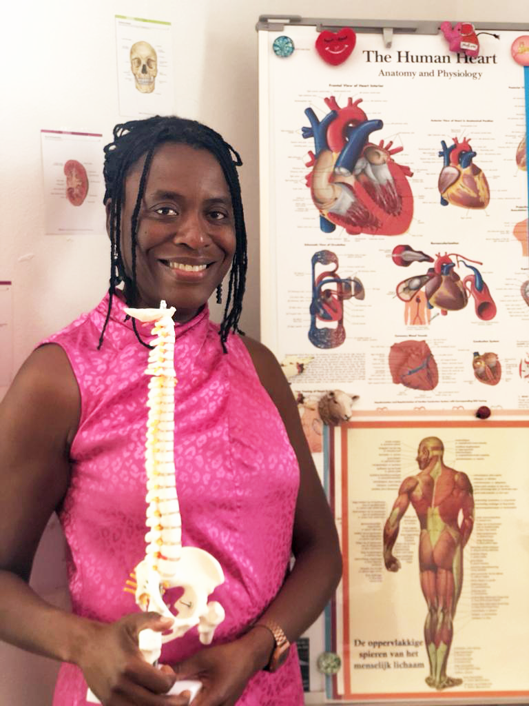 Vrouw in roze kleding die een menselijke ruggengraatmodel vasthoudt, staat voor anatomieposters van het menselijk lichaam en het hart in een klaslokaal of medisch instituut.