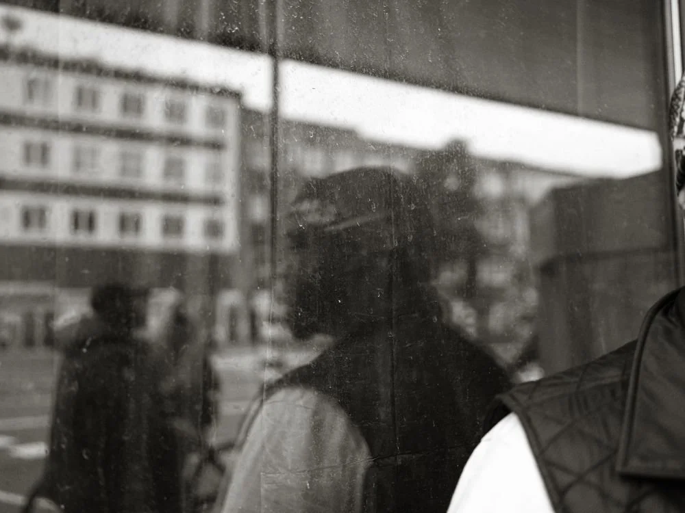 Mensen worden weerspiegeld in een glazen raam of deur, met een stedelijke achtergrond van gebouwen en mensen in de verte.