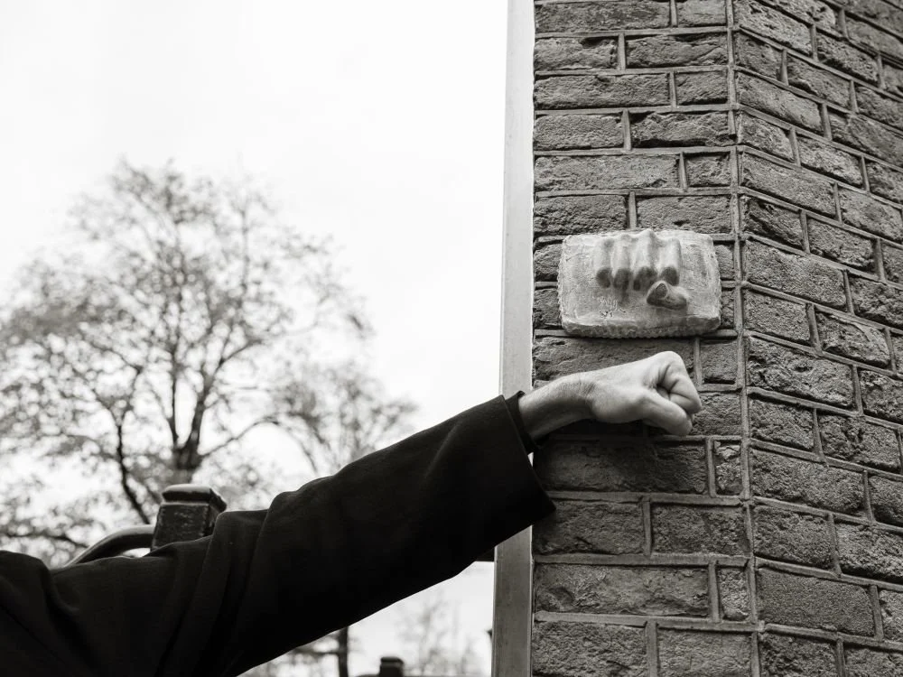 Een hand die een vuist vormt, uitsteekt naar een muur met een sculptuur van een hand en een voet, buiten op een dag met bewolkte hemel.