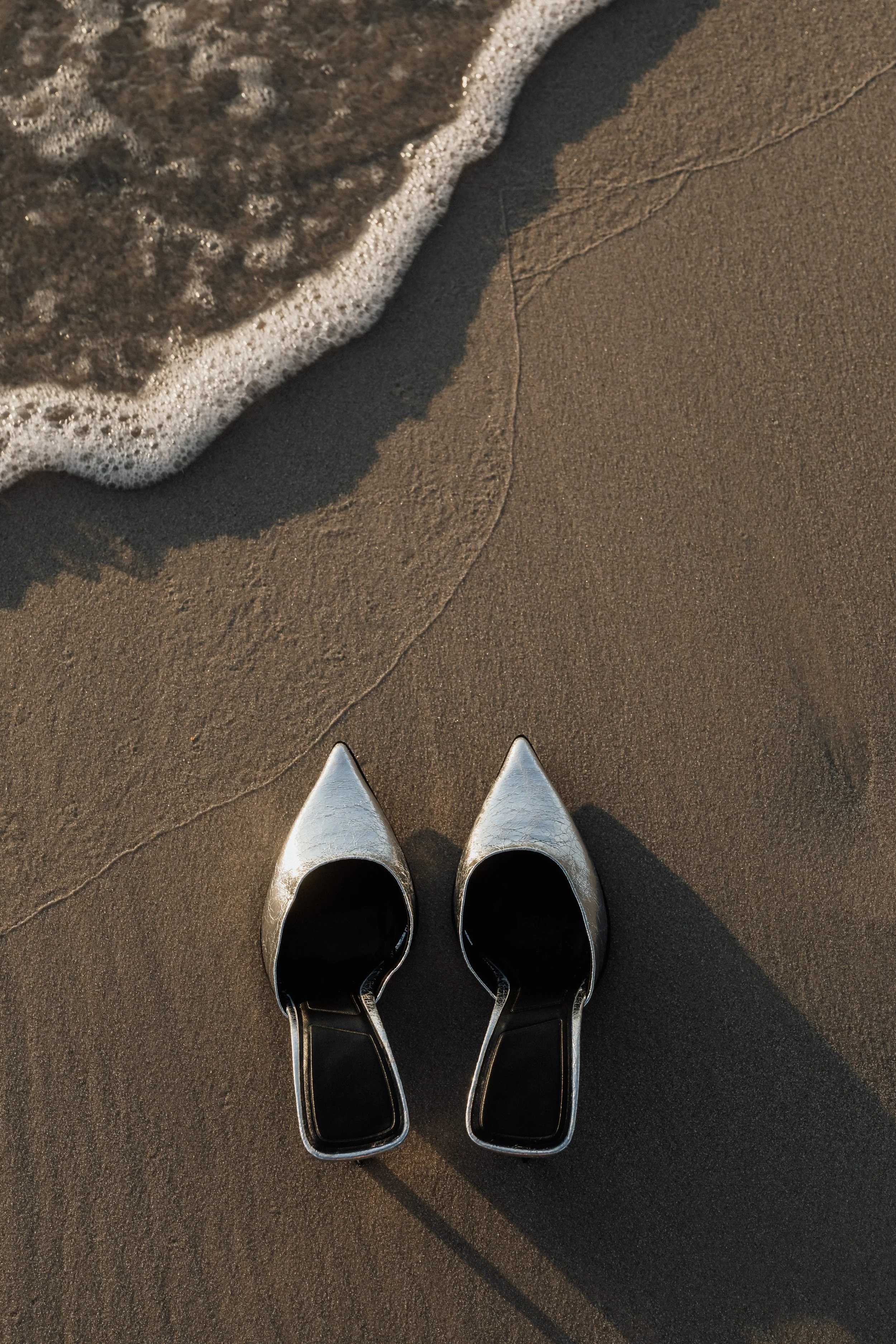 Twee paar zilveren hoge hakken op het zandstrand met een golf die nadert.