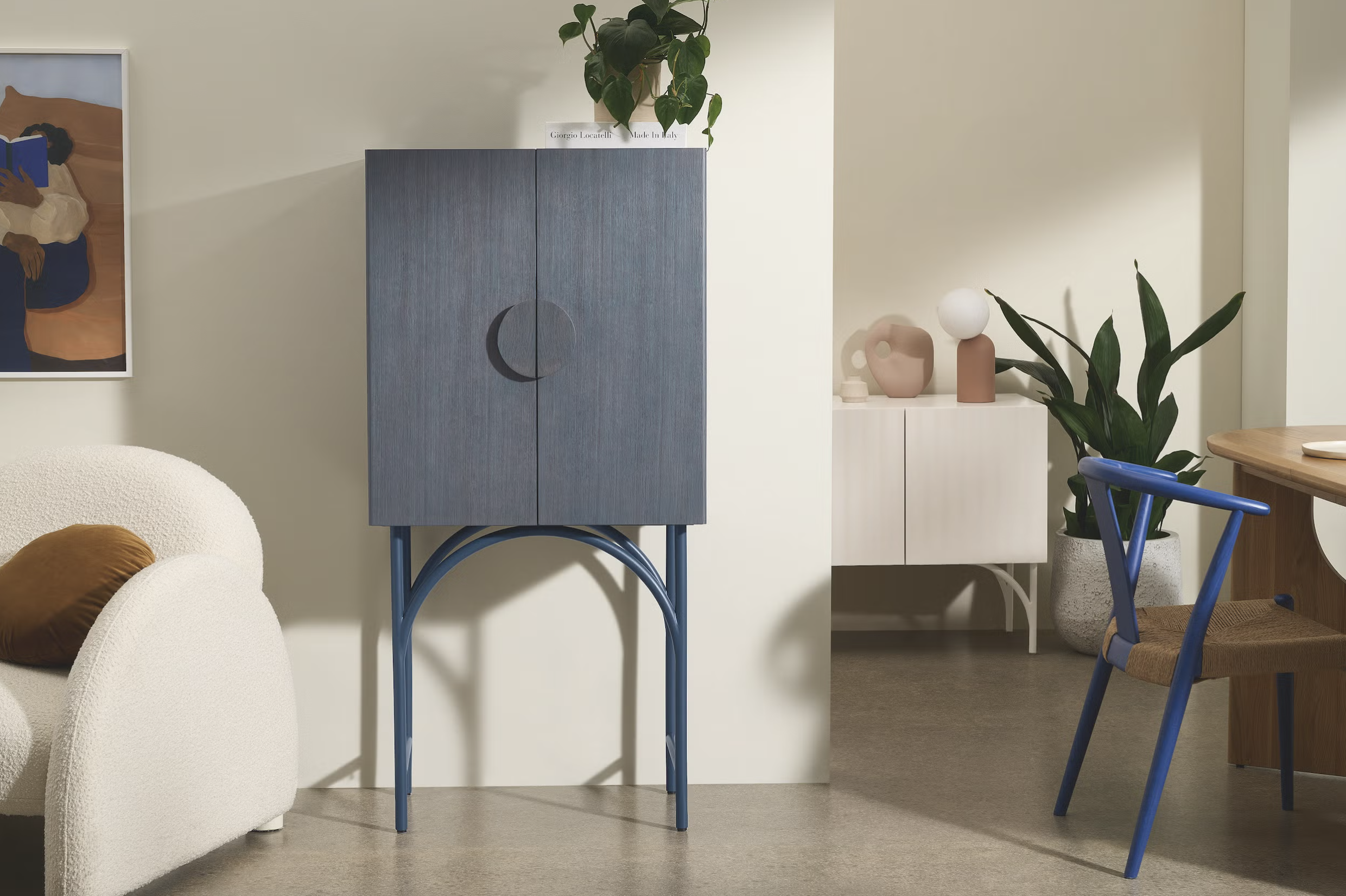 Interieur van een woonkamer met een witte bank, een bruin kussen, een blauw houten kast op blauwe metalen poten, een plantenbak met groene bladeren, een witte en beige sideboard met vazen en een grote plant, en een tafel met een blauwe stoel.