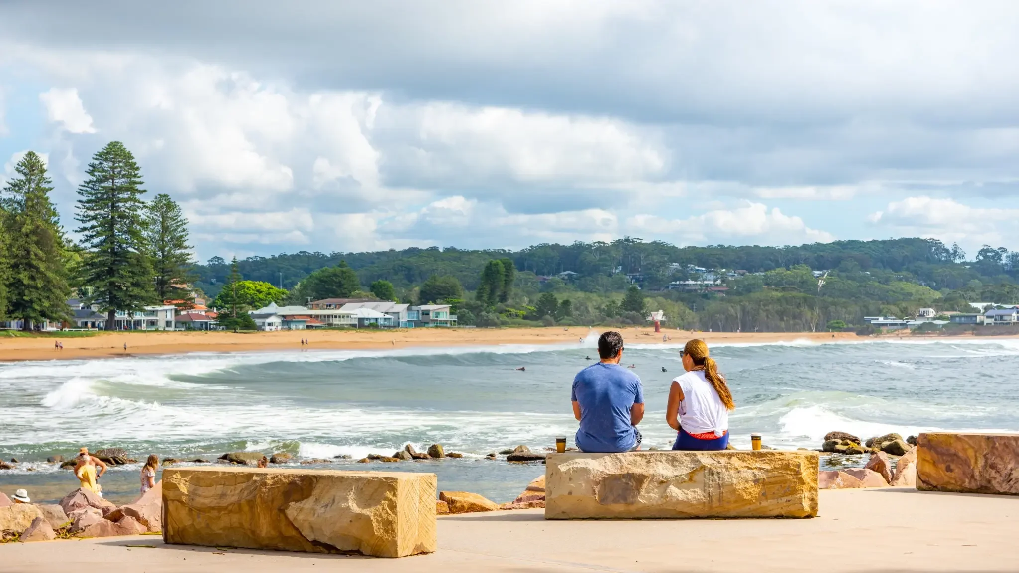 People sitting at Avoca Beach promenade looking out over the waves on a cloudy day