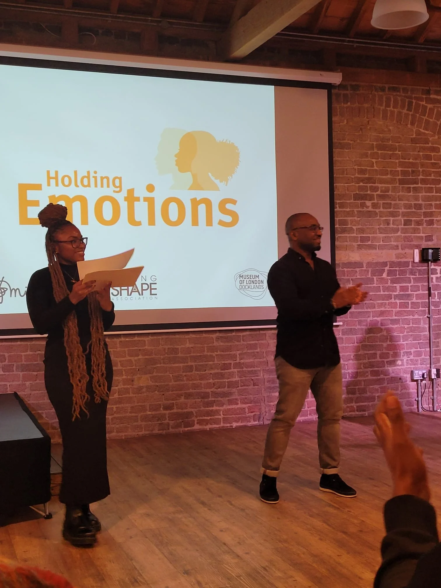 A woman with long braided hair and glasses holding papers, and a man with glasses clapping, are standing in front of a presentation screen that reads 'Holding Emotions' with silhouettes of a woman's profile in various shades of yellow. The setting appears to be an indoor event or workshop in a room with exposed brick walls.