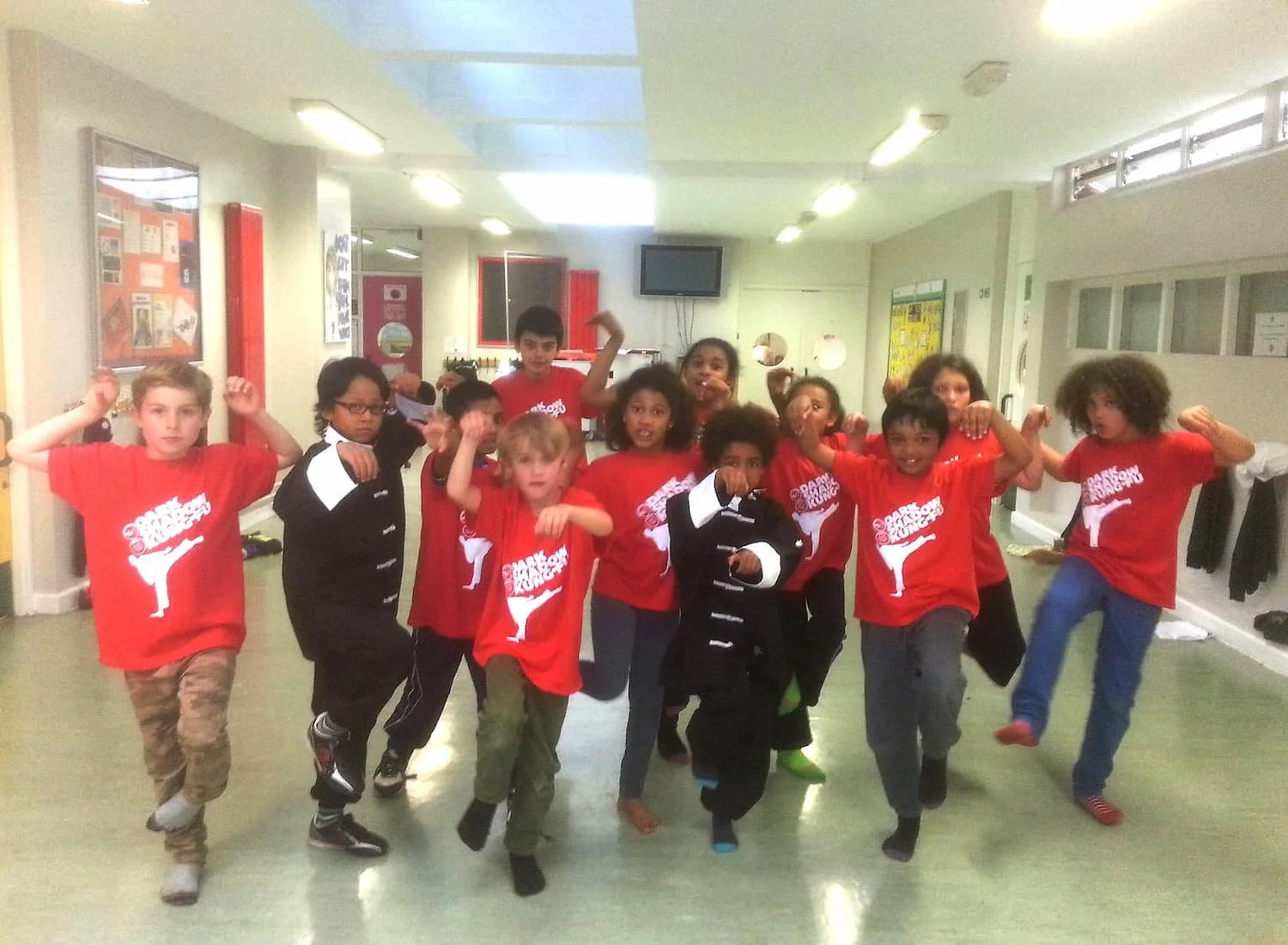 Group of children doing martial arts poses indoors, some wearing red shirts with white text and logo, others in black and white outfits.