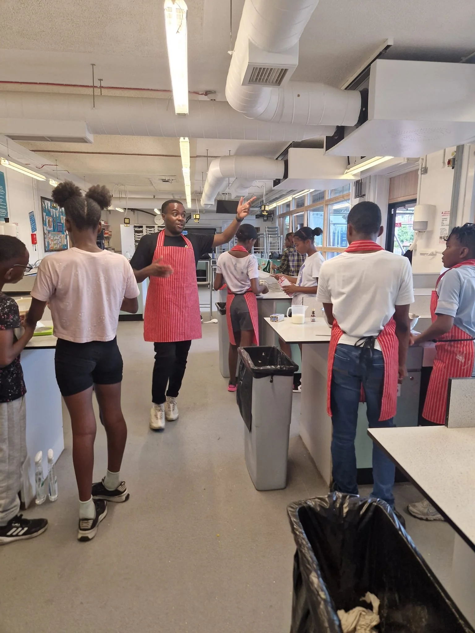 Instructor leading a cooking class with students in a commercial kitchen, wearing aprons, some holding utensils.