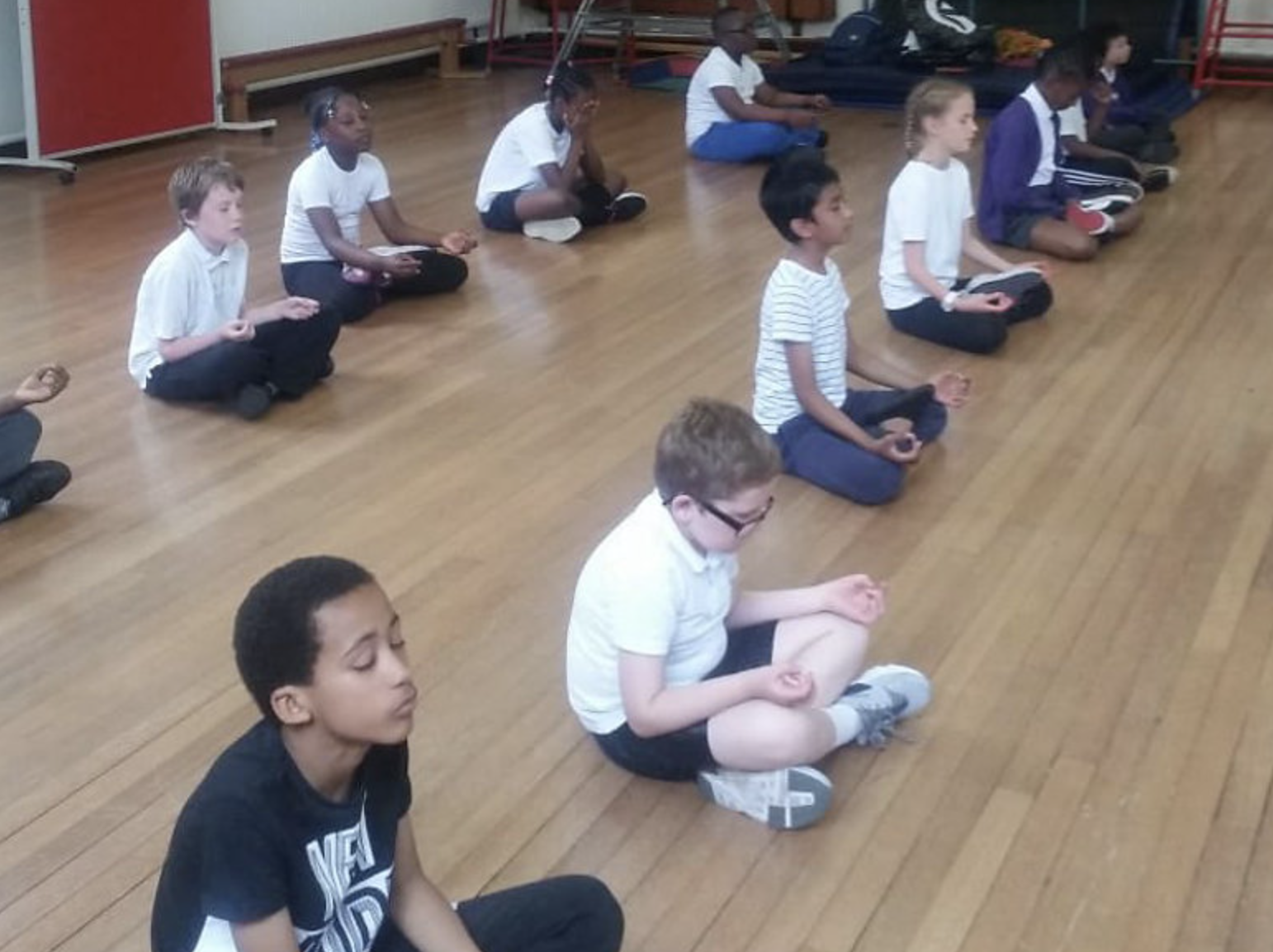 Children seated on the hardwood floor in a meditation or mindfulness class, sitting cross-legged with eyes closed, in a school or community center.