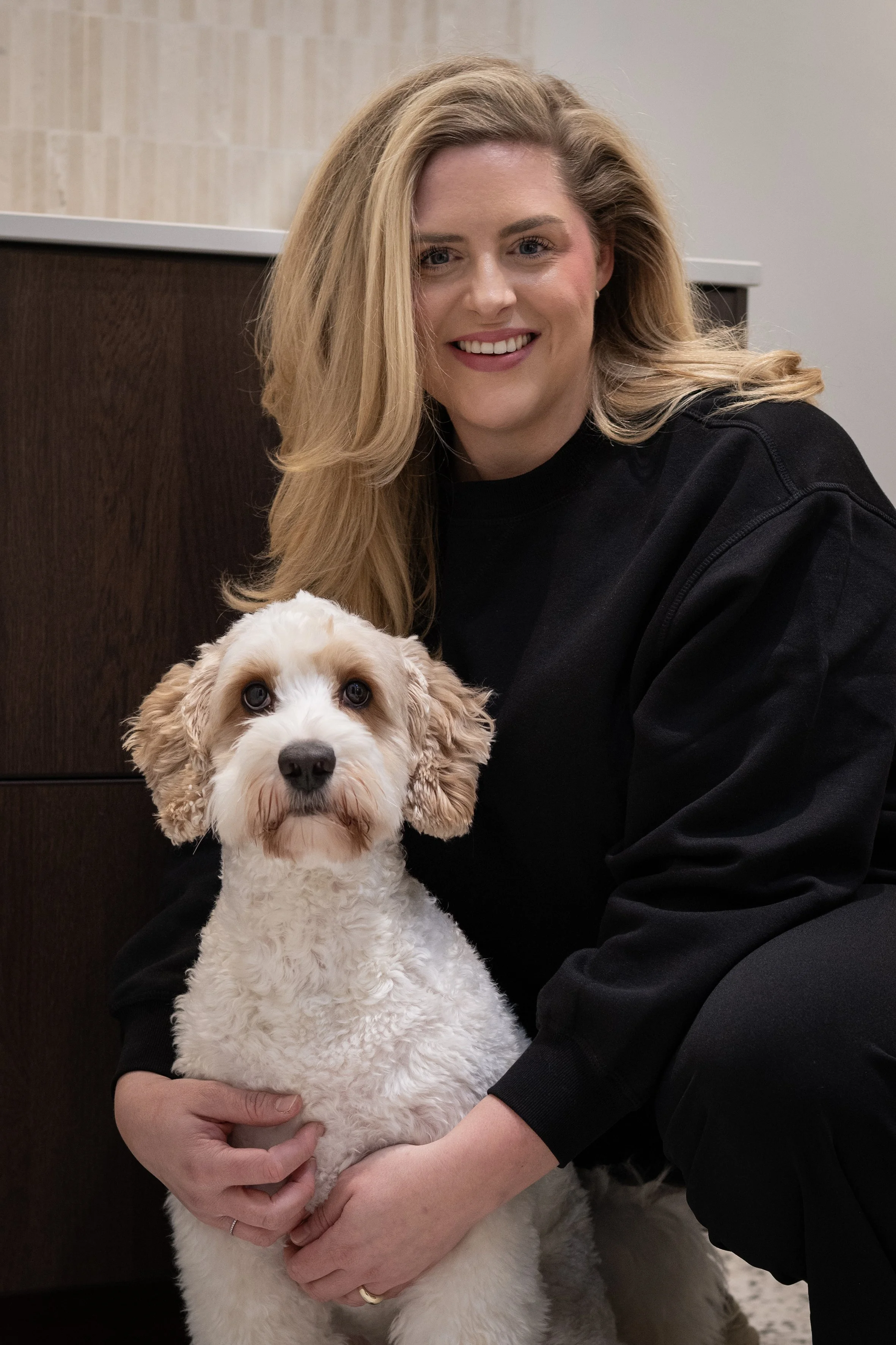 A smiling woman with long blonde hair wearing a black sweatshirt holding a white and brown curly-coated dog in an indoor setting.