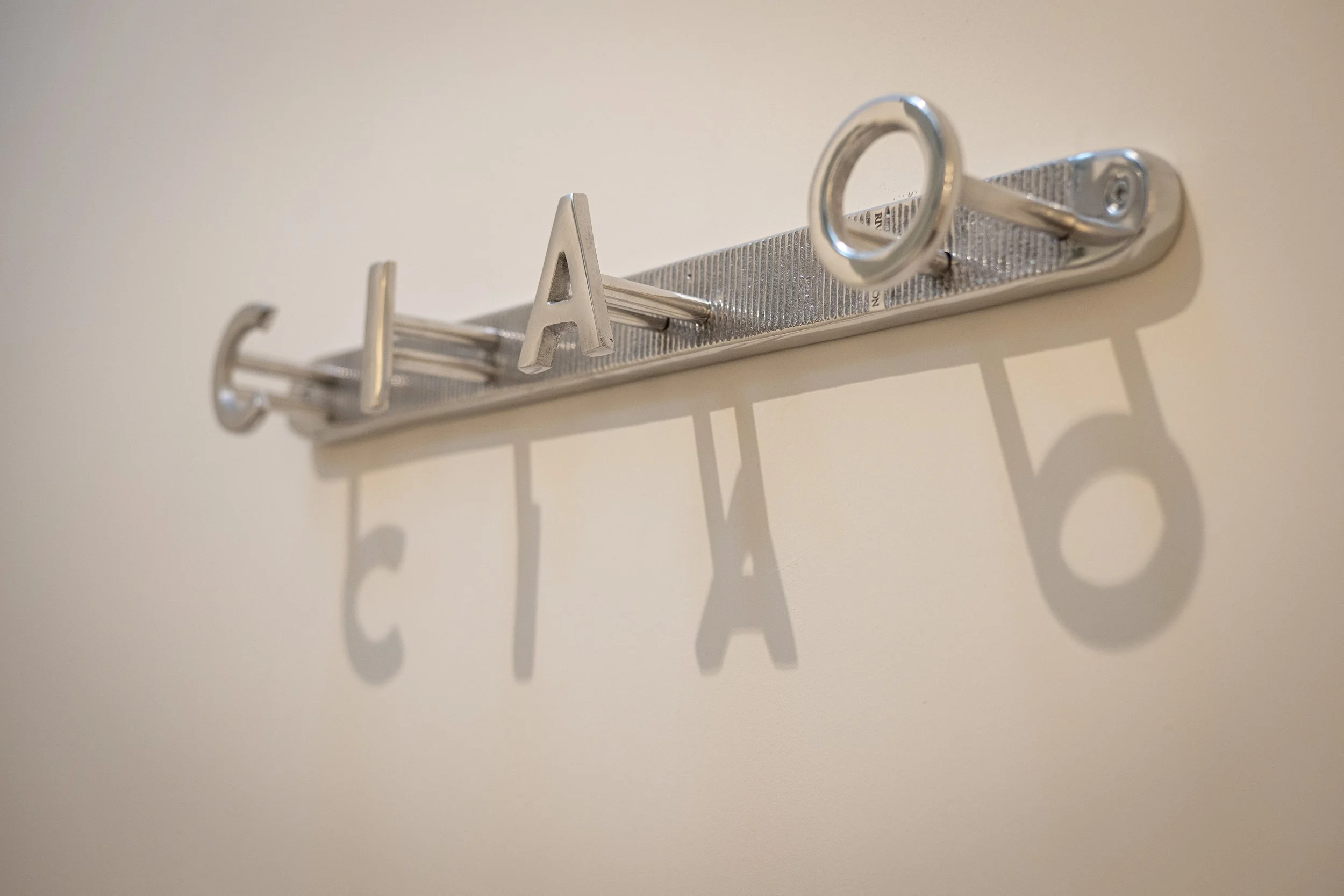 Wall-mounted metallic coat rack with large 3D letters spelling 'CATA' casting shadows on the wall.