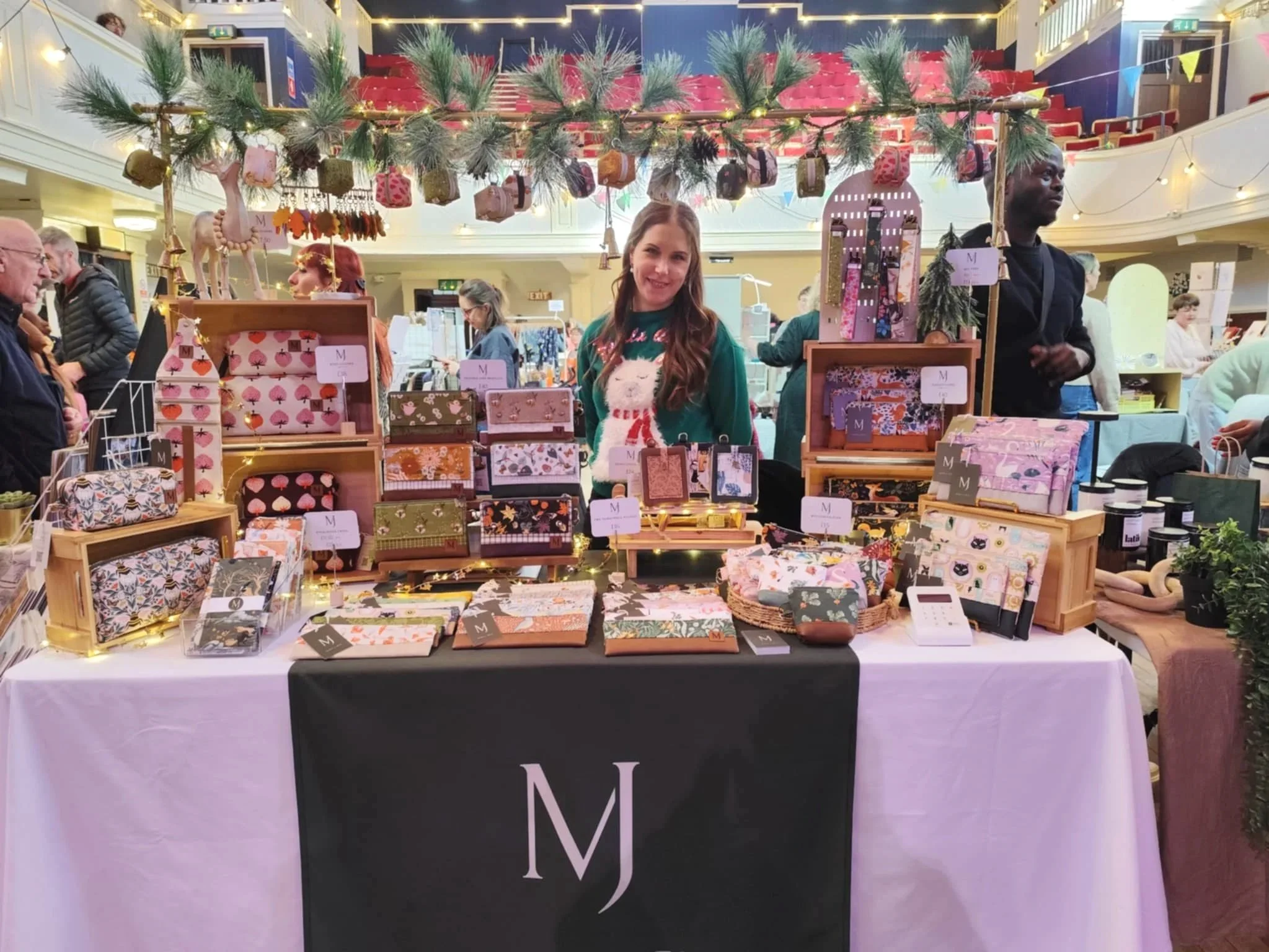A woman standing behind a table at a craft fair booth decorated with Christmas greenery and lights, selling colorful patterned wallets and accessories. Several people browse around in the background.