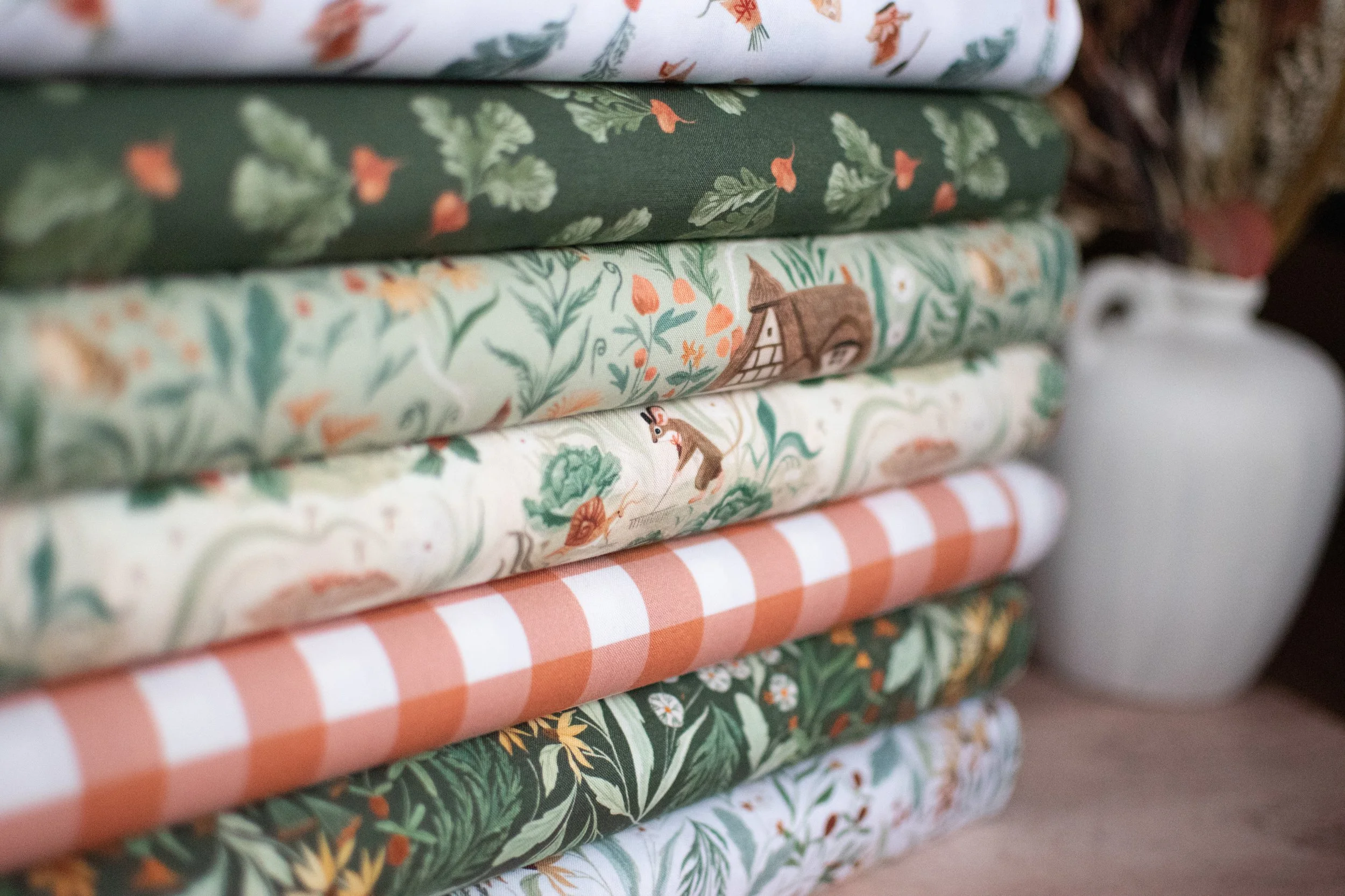 Stack of folded fabric with various nature-inspired and checkered patterns, with a white jug in the background.