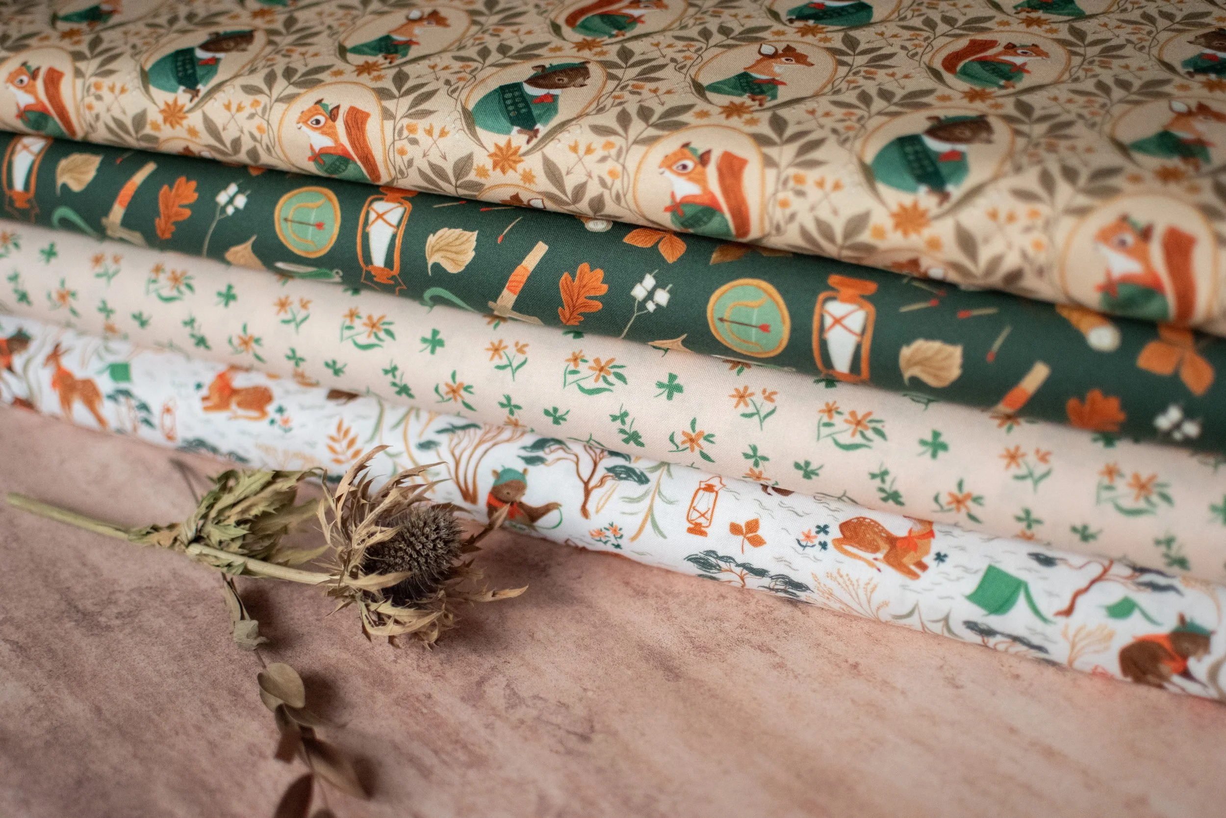 Stacked Christmas-themed fabrics with woodland animals and nature patterns, placed on a textured surface with a dried flower arrangement.
