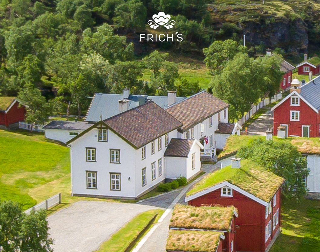 Dette er Kongsvoll gjestegård. Det er et landskap med høye, grønne trær og flere tradisjonelle norske hus, noen med hvit maling og andre med rød maling, alle med ulike takmaterialer. Veier og gresskledde områder omgir husene.