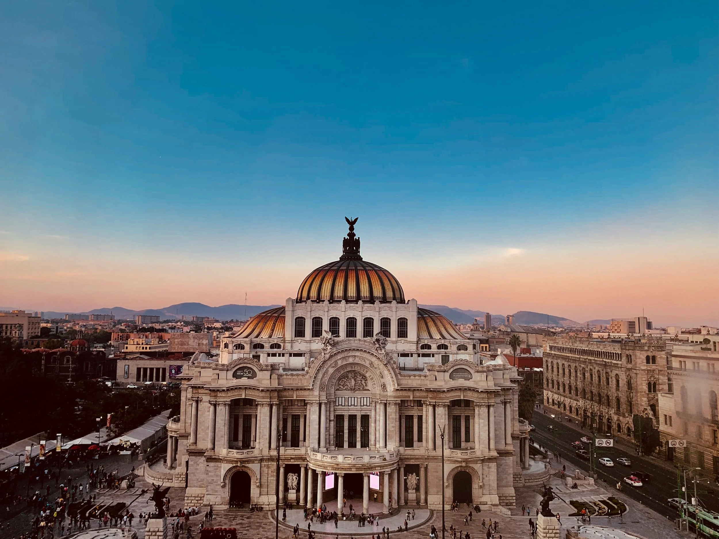 The Palace of Fine Arts in Mexico City illuminated during sunset, with a clear sky and mountains in the background.