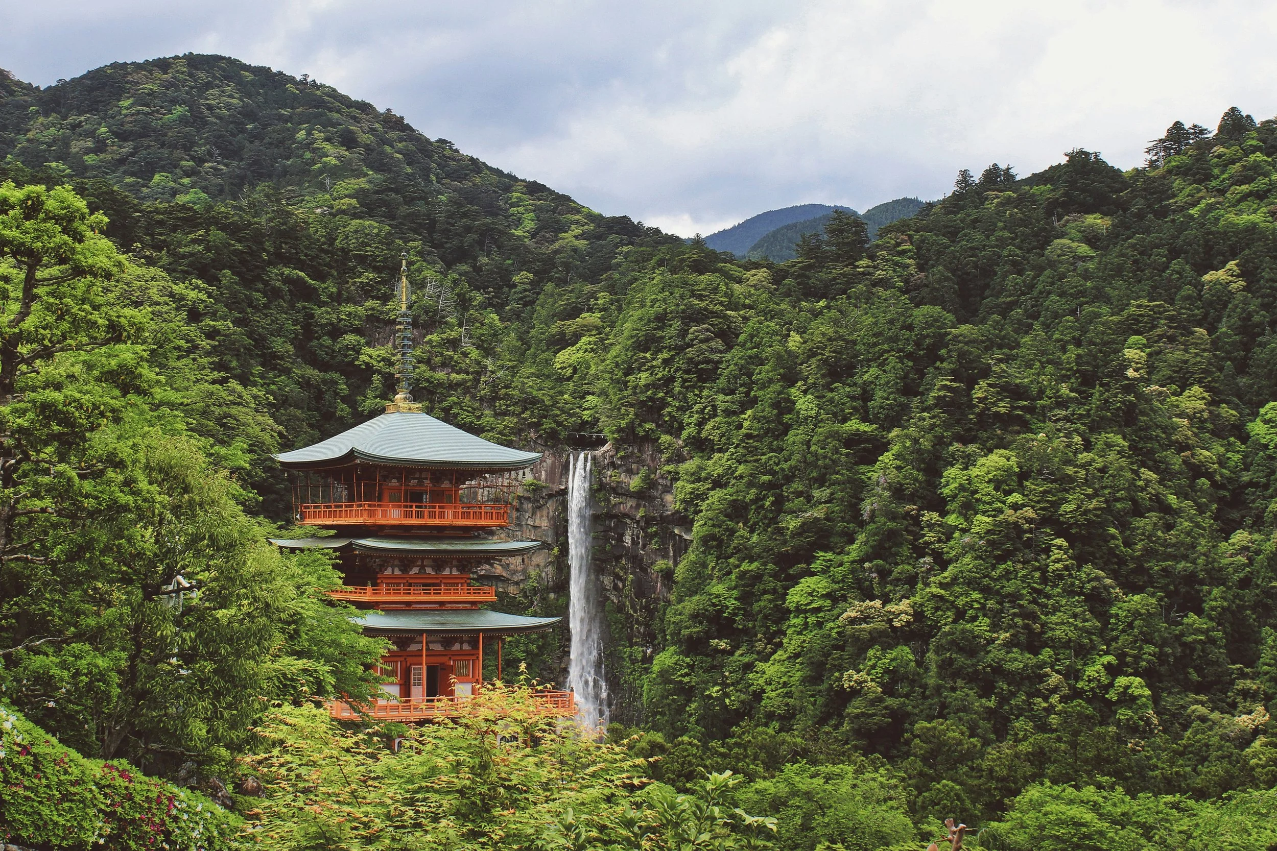 A traditional Japanese pagoda with a waterfall behind it, surrounded by lush green trees and mountains.