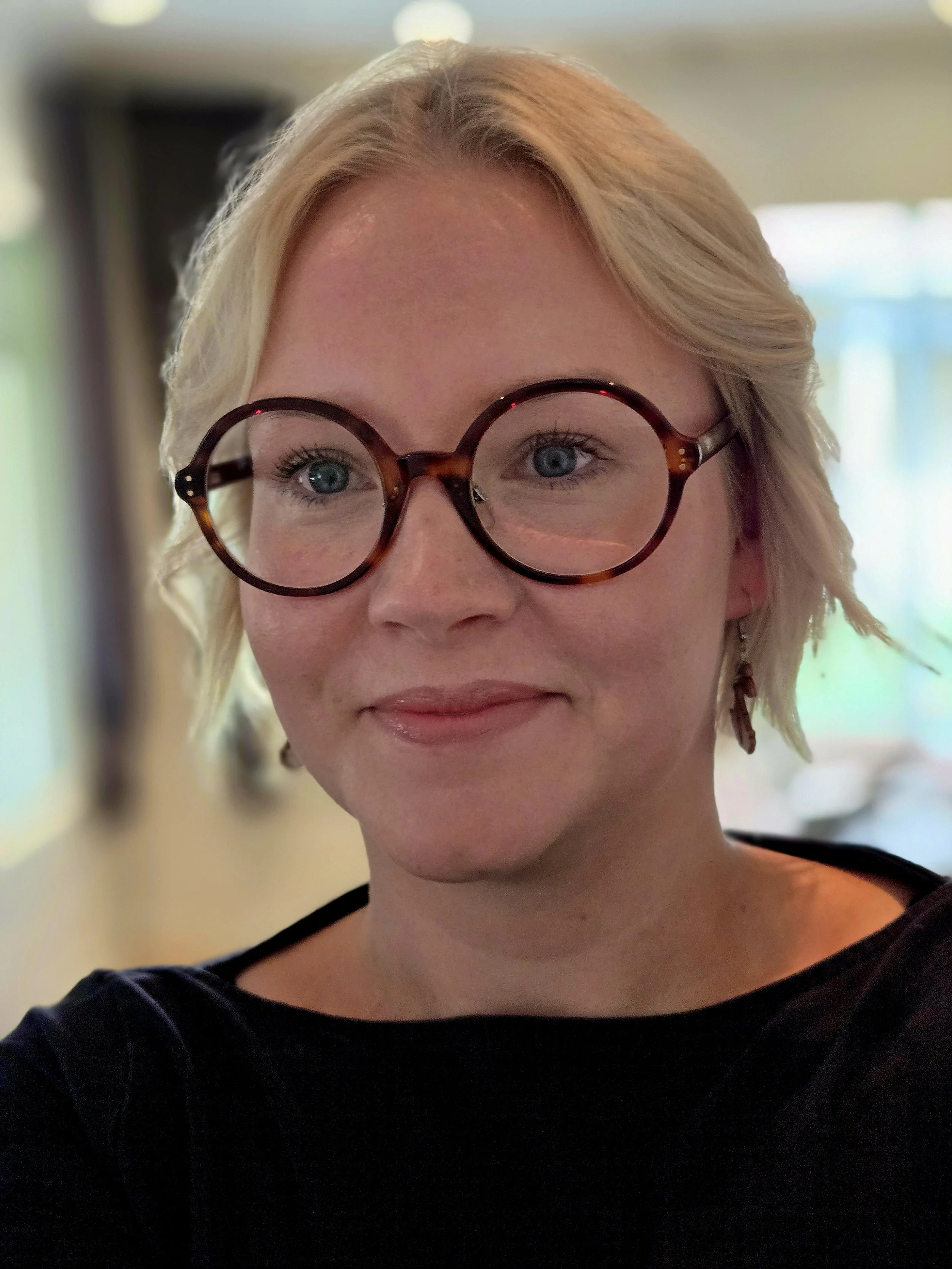 Naisella ihmisellä on vaaleat hiukset ja paksut pyöreät ruskeat silmälasit. Hän on hymyilevä ja katsoo hieman kameran yläpuolelle. Taustalla on epätarkka sisätila.