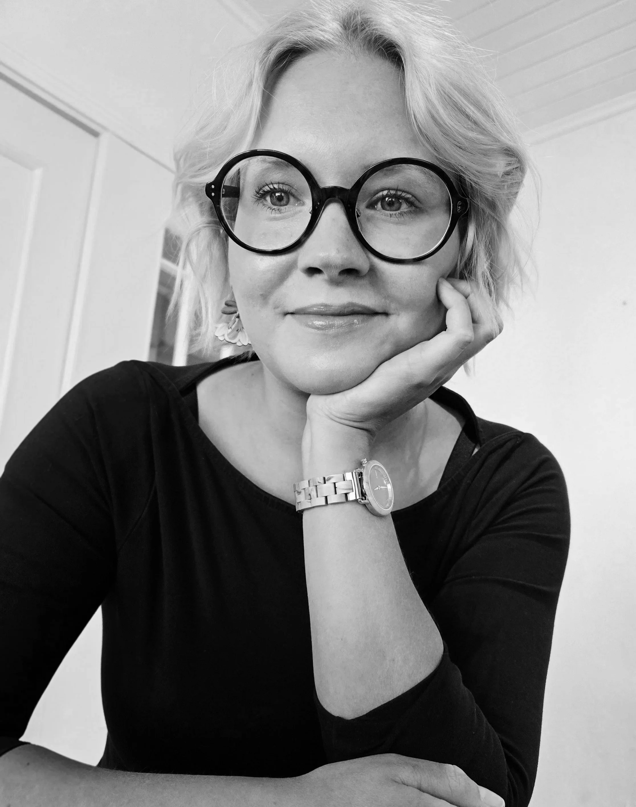 Vaalea nainen hymyilee, nojaa leukaansa käteen ja katsoo kameraan