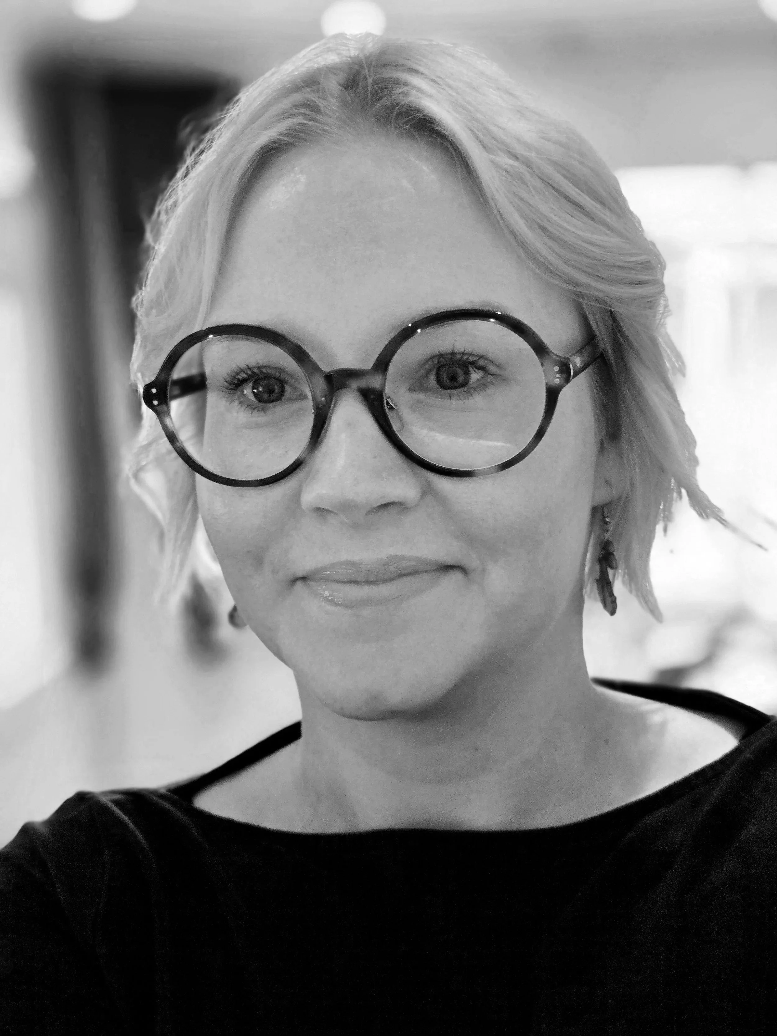 Nainen pyöreissä kyhyksissä, pyöreät isot silmälasit, hymyilevä ilme.